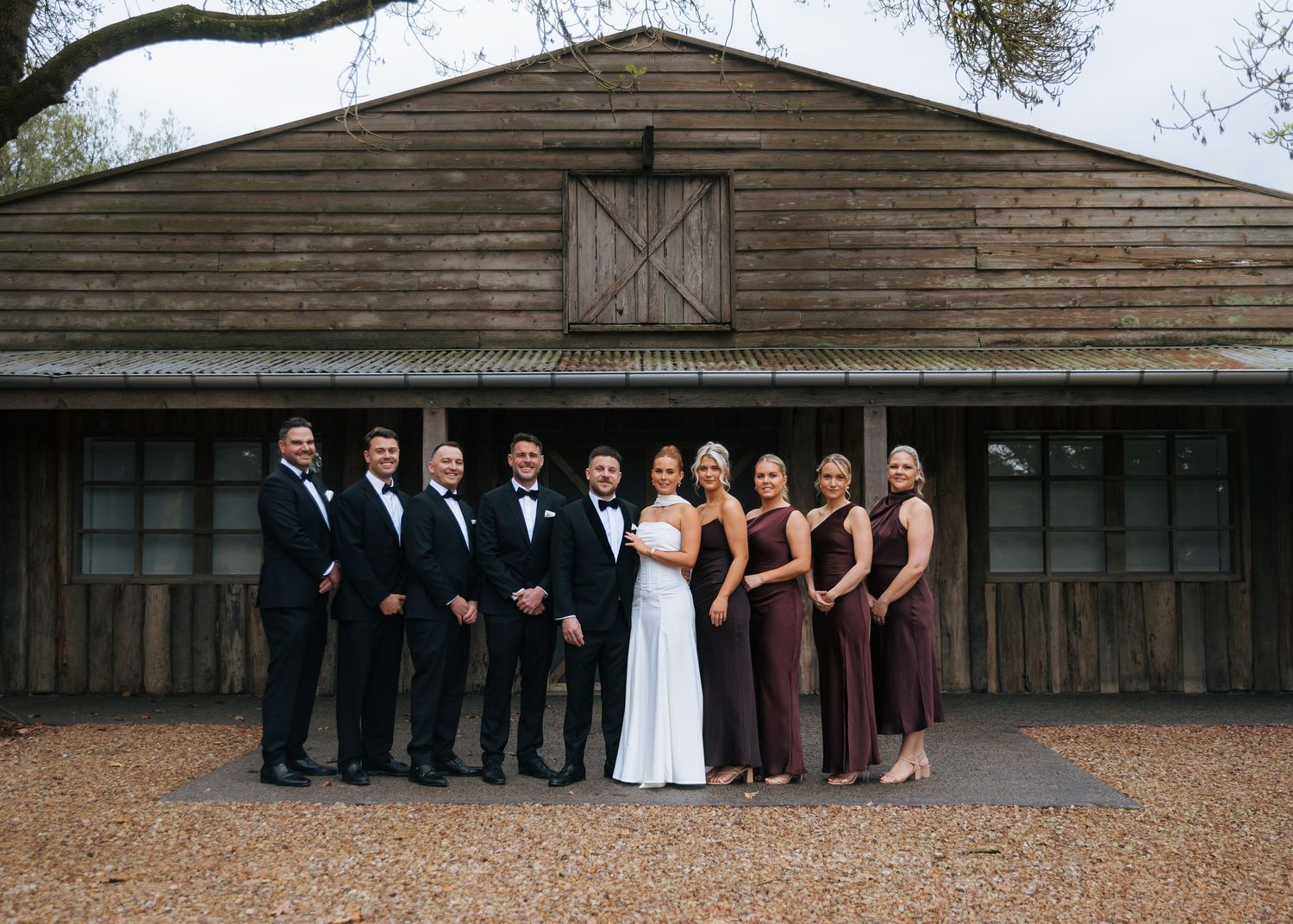 Wedding party poses in front of rustic barn venue, groomsmen in tuxedos and bridesmaids in burgundy dresses.