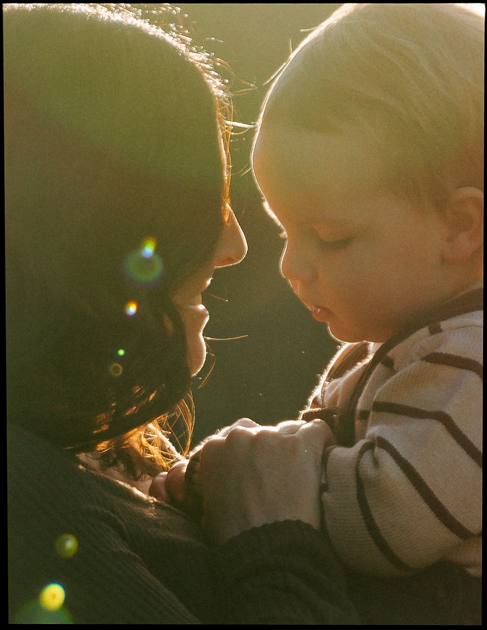Tender close-up of mother and toddler touching noses in warm golden sunlight outdoors.