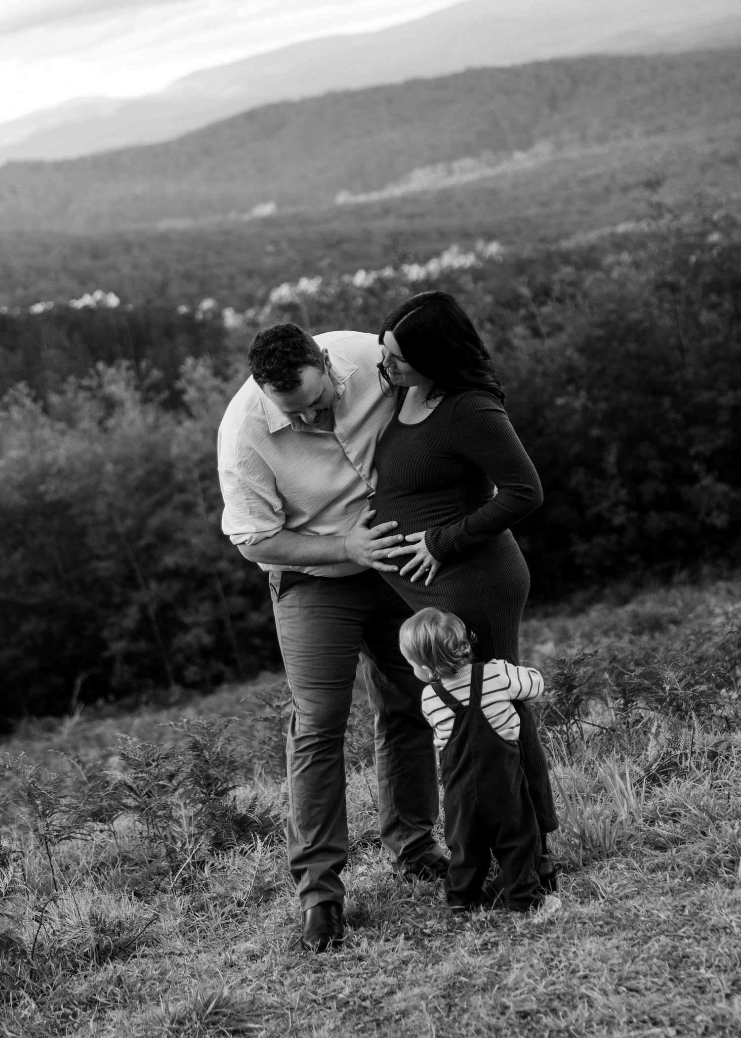 Black and white photo of couple with toddler embracing in a field with mountain backdrop.