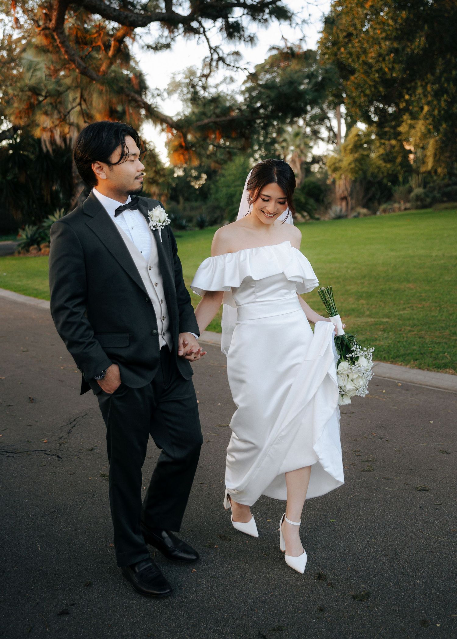 Bride lifting her white satin gown while walking hand in hand with groom in black tuxedo along a park path.