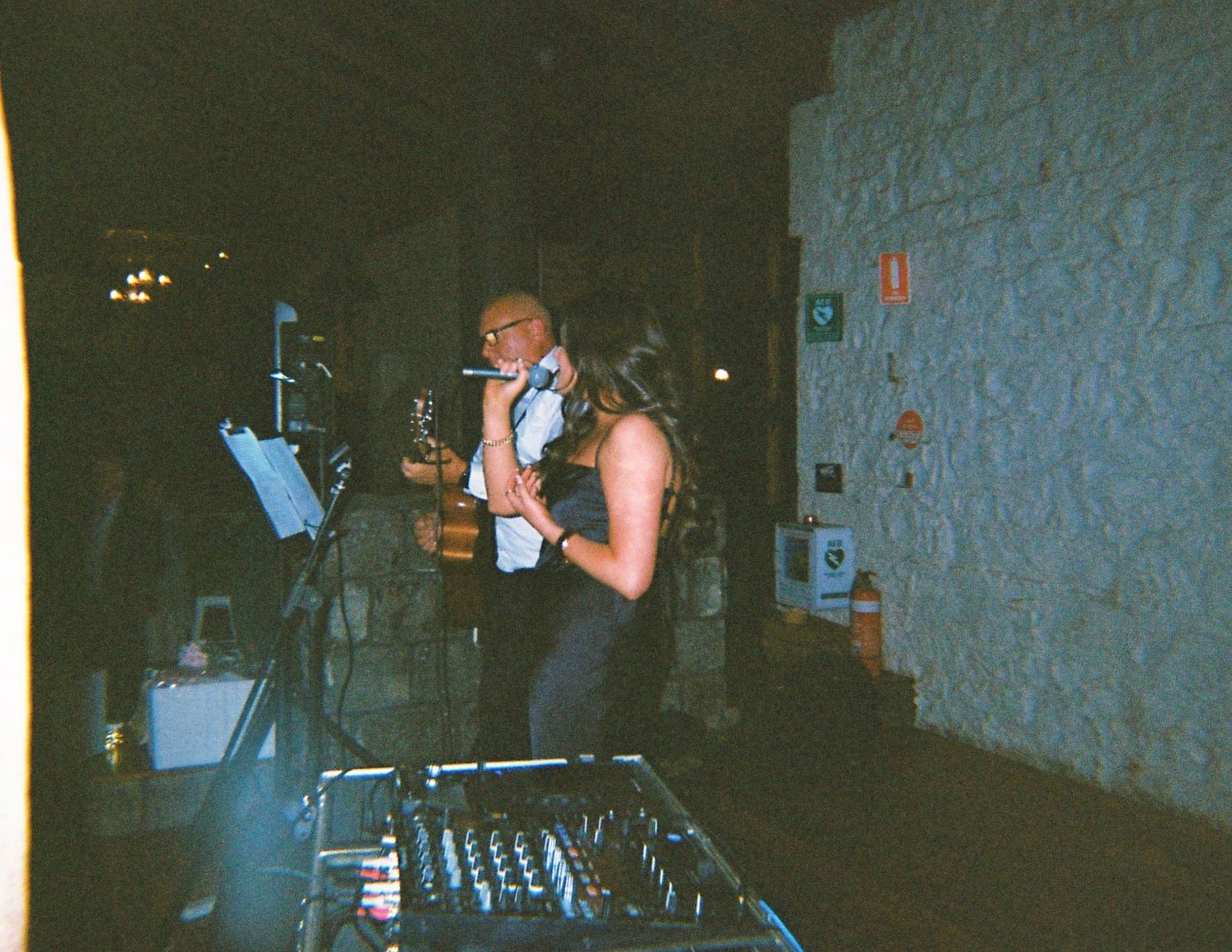 A musician plays saxophone on stage at a dimly lit wedding reception with a DJ mixing console in the foreground.