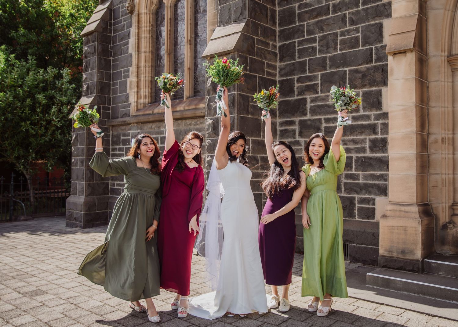 Bride and four bridesmaids in colorful dresses joyfully raise bouquets outside a stone church.