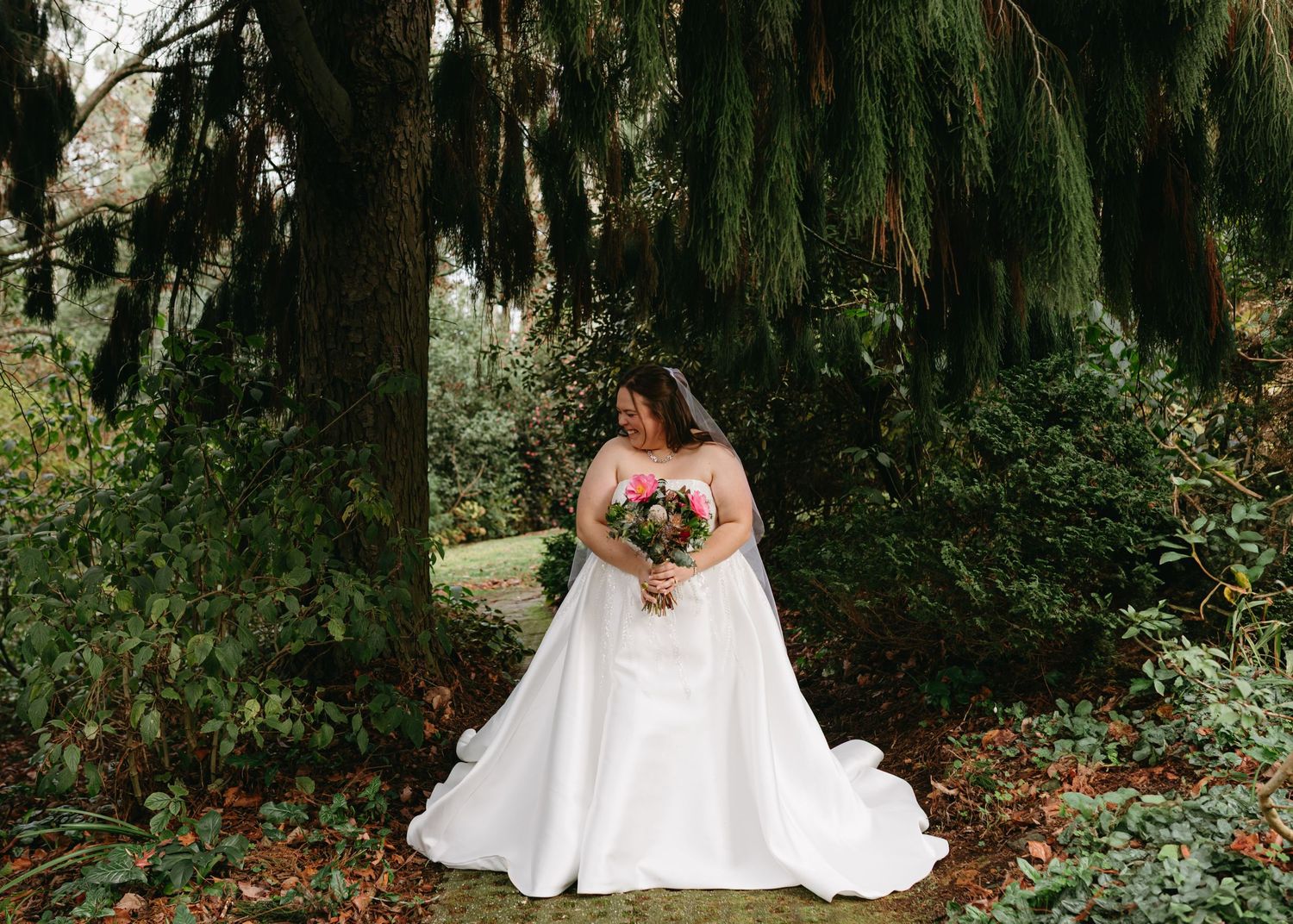 Bride in a white ballgown holding a floral bouquet surrounded by tall green trees.