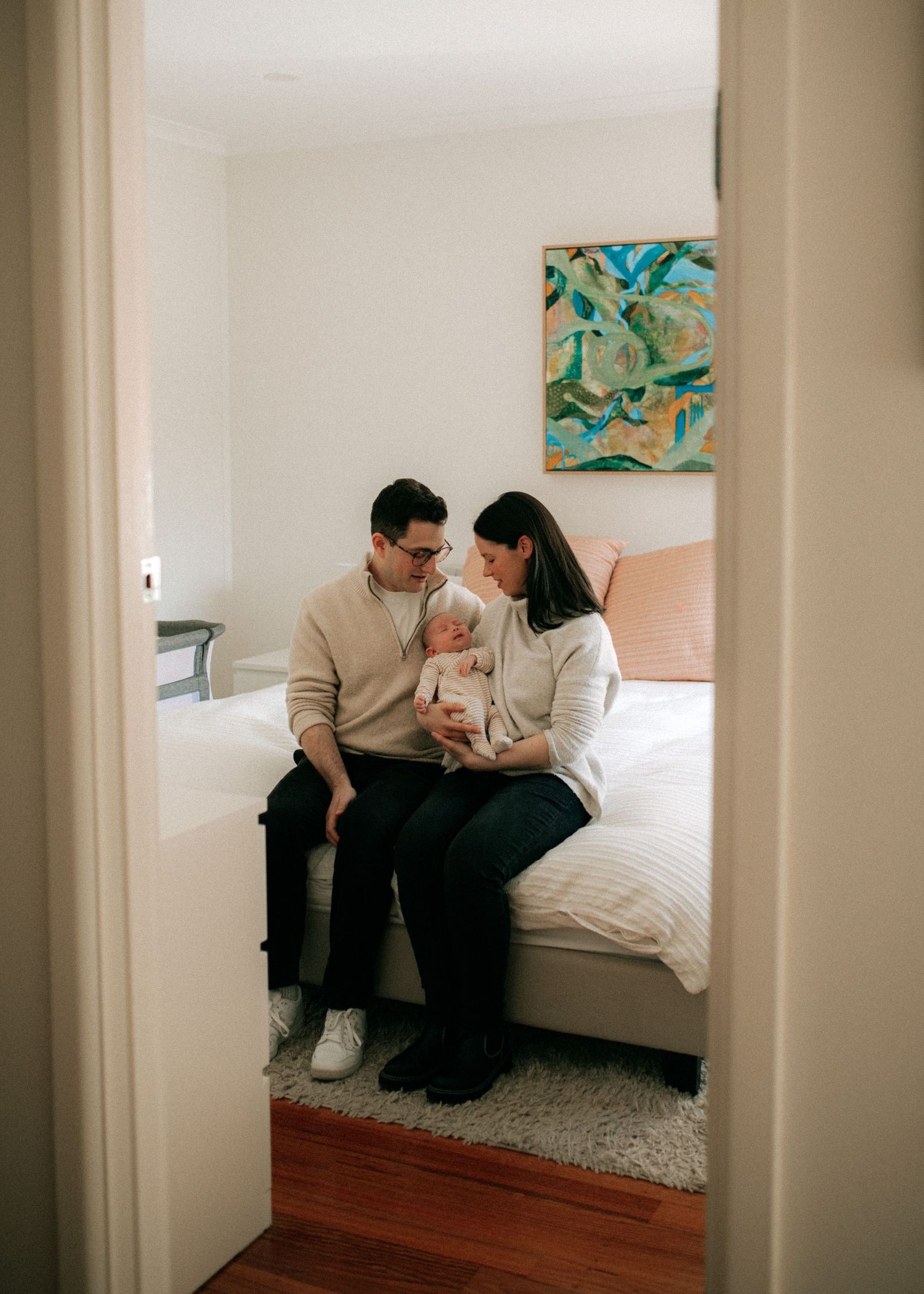 New parents sit on a bed holding their newborn baby, viewed through a doorway in a cozy bedroom.
