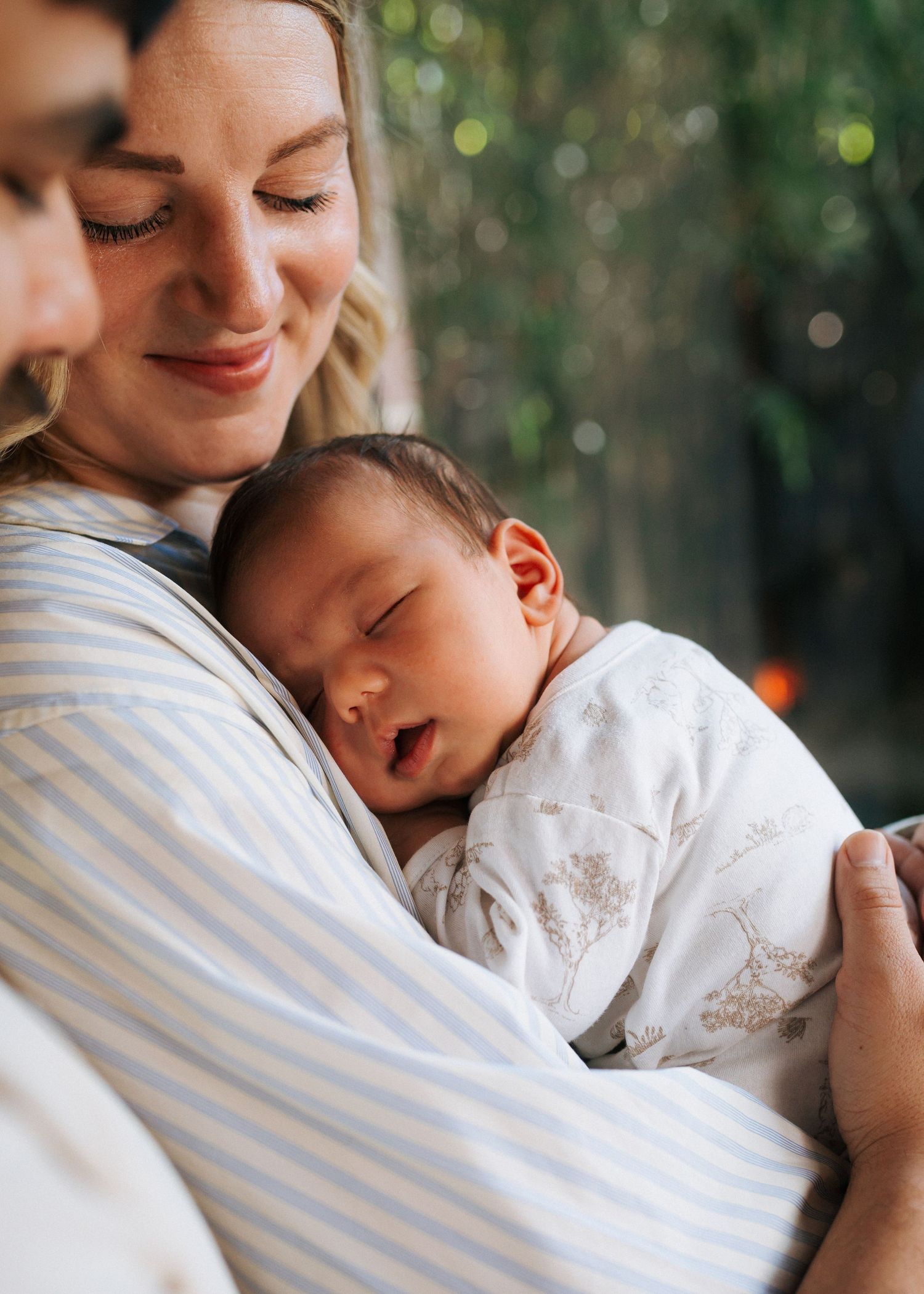 A smiling woman in a striped shirt gently cradles a sleeping newborn baby in a white floral outfit against a soft, blurred green background.