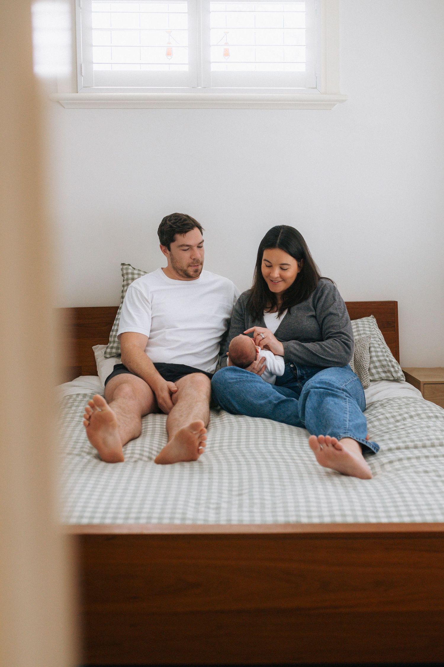 Smiling couple sits barefoot on a wooden bed with gingham bedding, cradling their sleeping newborn baby in a bright, cozy bedroom.