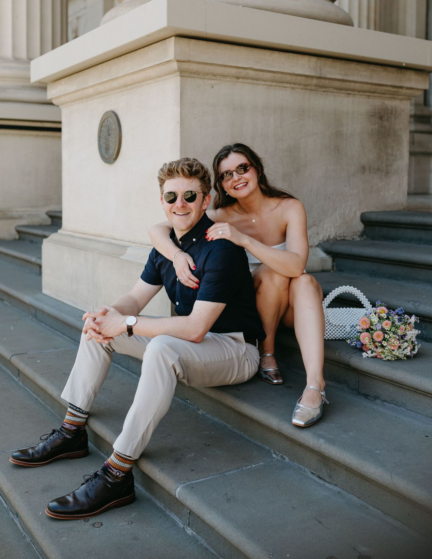 Smiling couple sitting on stone steps of a neoclassical building, relaxing together with a floral bouquet nearby.