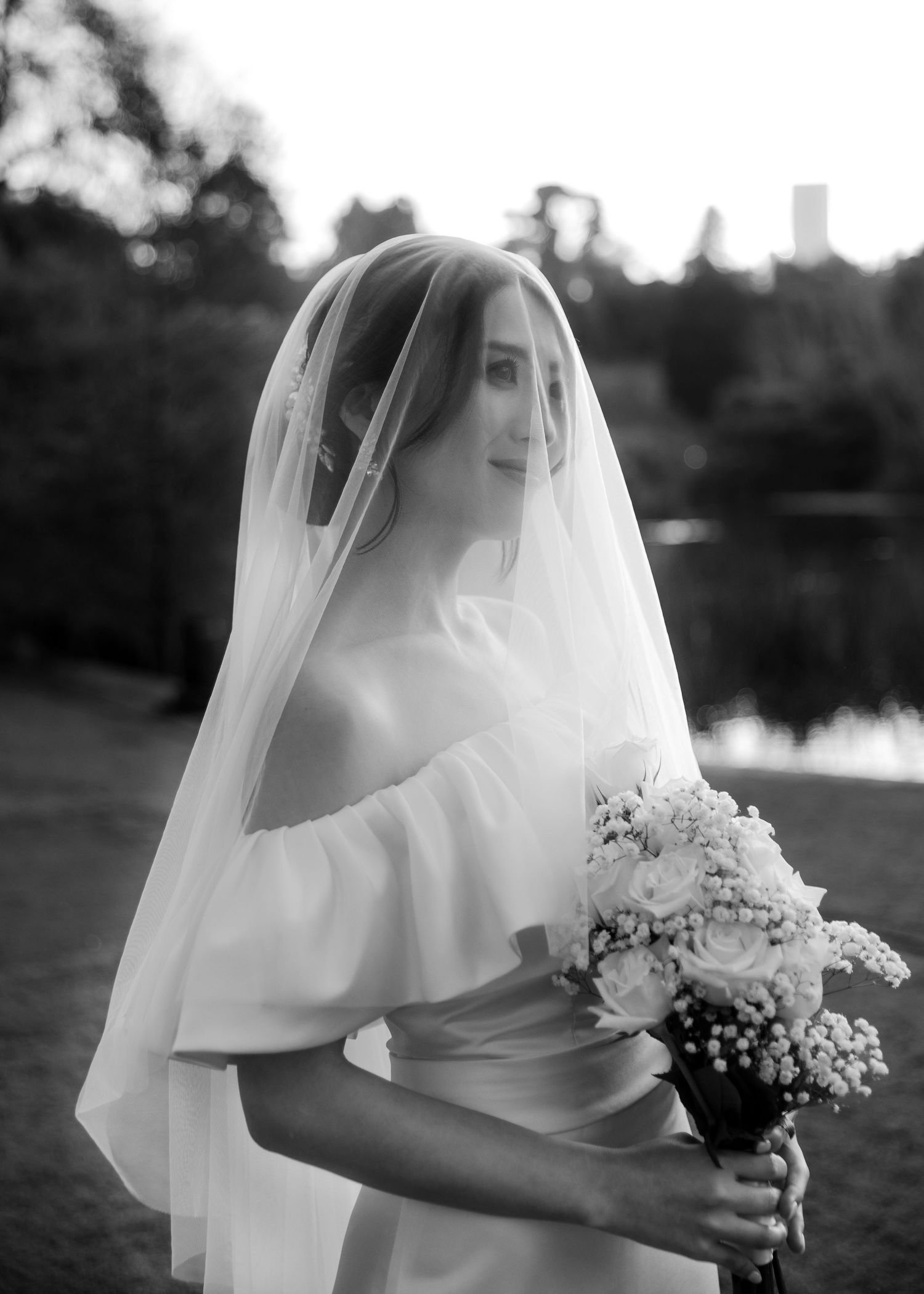 Elegant bride in off-shoulder gown holding white bouquet under flowing veil at sunset.