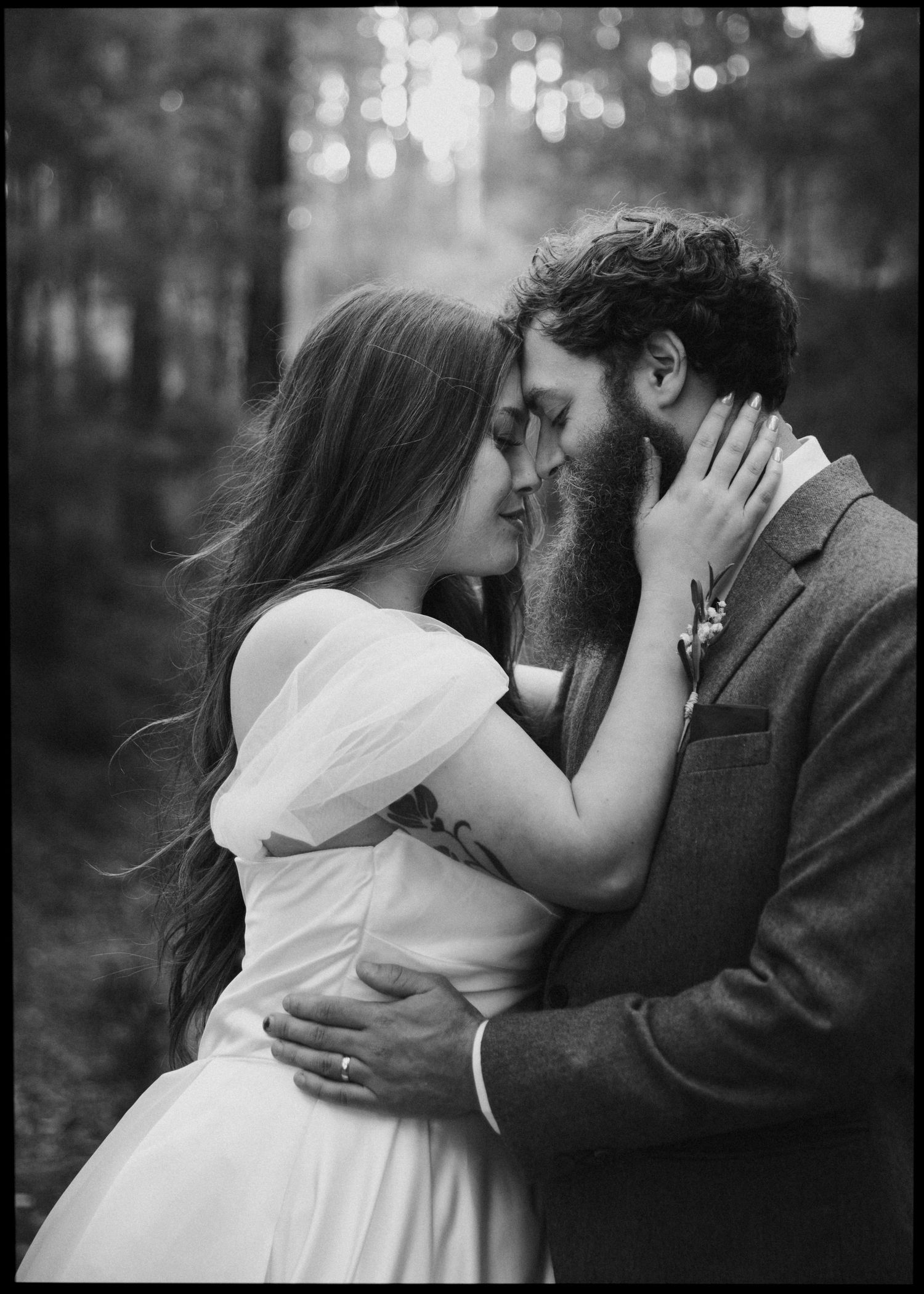 Black and white romantic portrait of a bride and bearded groom nearly kissing in a misty forest.