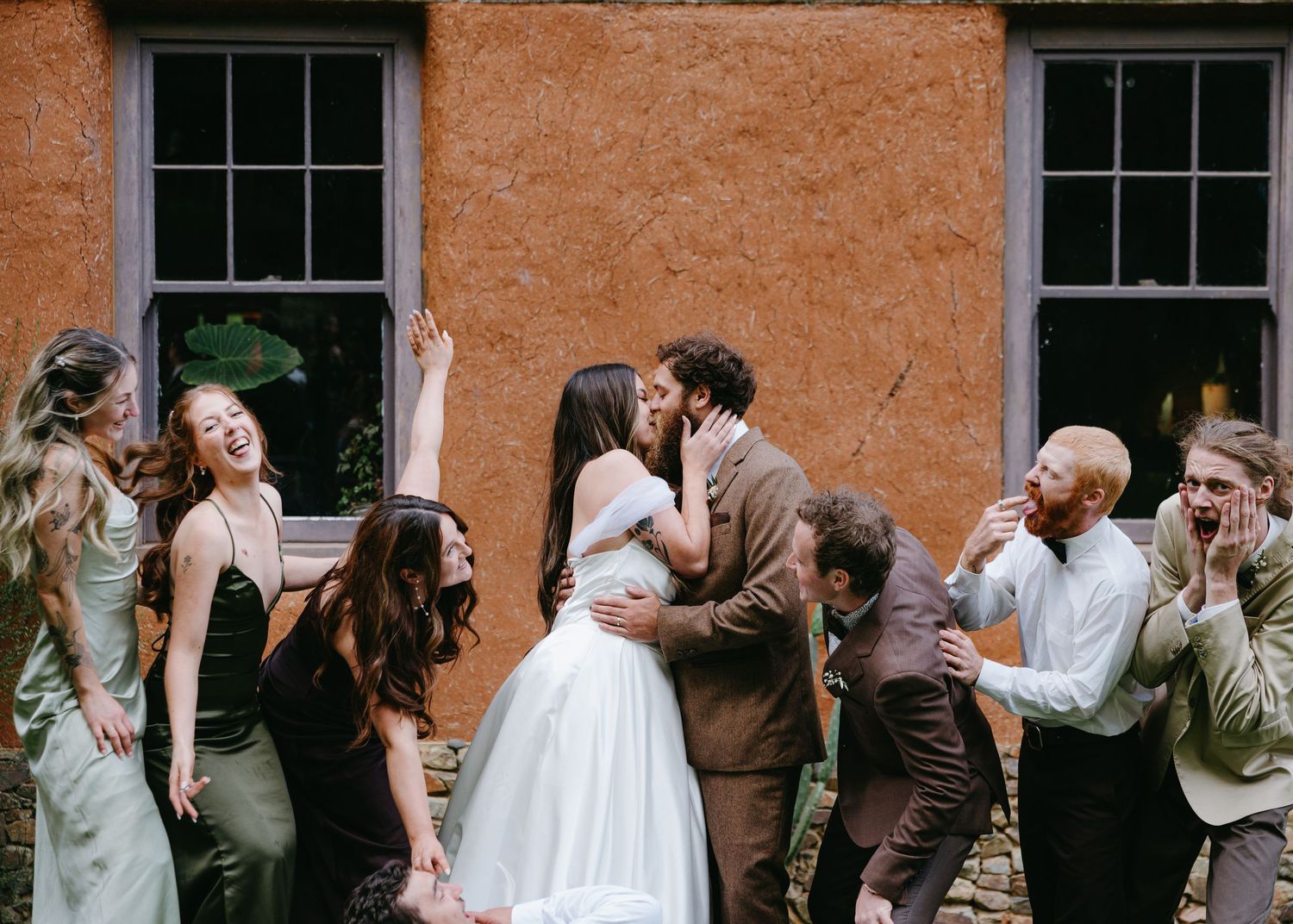 Bride and groom sharing a kiss surrounded by cheering bridesmaids and groomsmen against a terracotta wall.