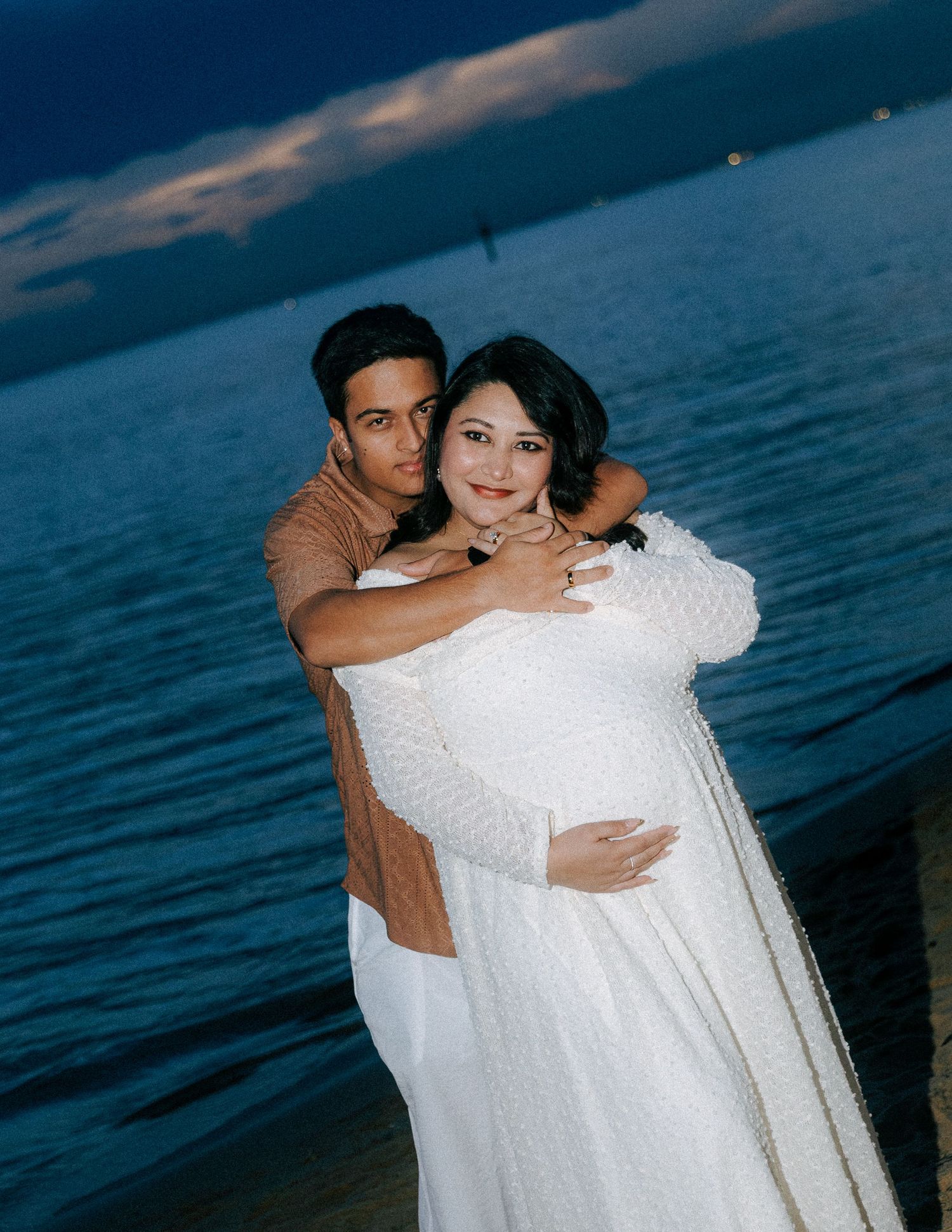 Couple sharing a tender maternity photo shoot at dusk by the water, woman in white lace gown cradling baby bump.