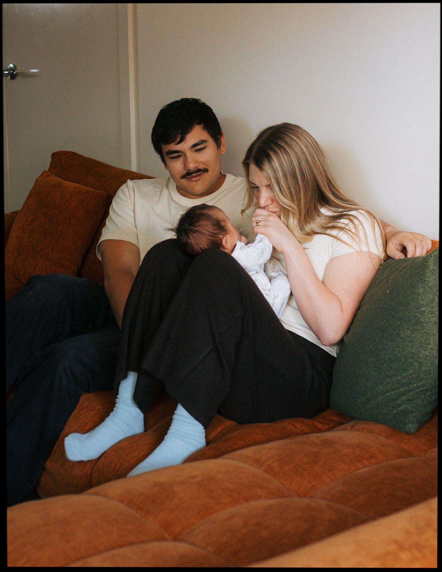 A couple sits close on a rust-orange velvet couch, gently cradling and kissing their sleeping newborn baby in a cozy, sunlit living room.