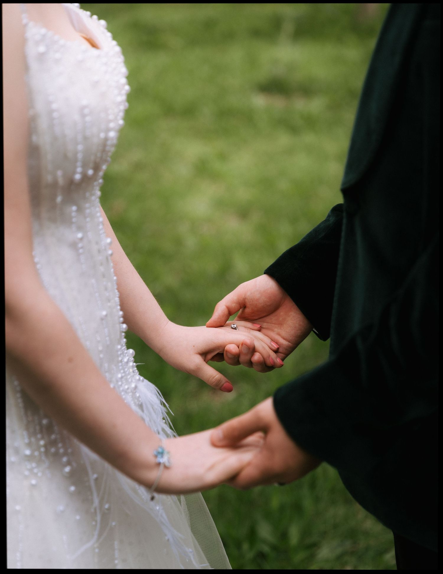 Bride in a beaded white wedding gown holds hands with a person in a dark robe during an outdoor ceremony on a green lawn.
