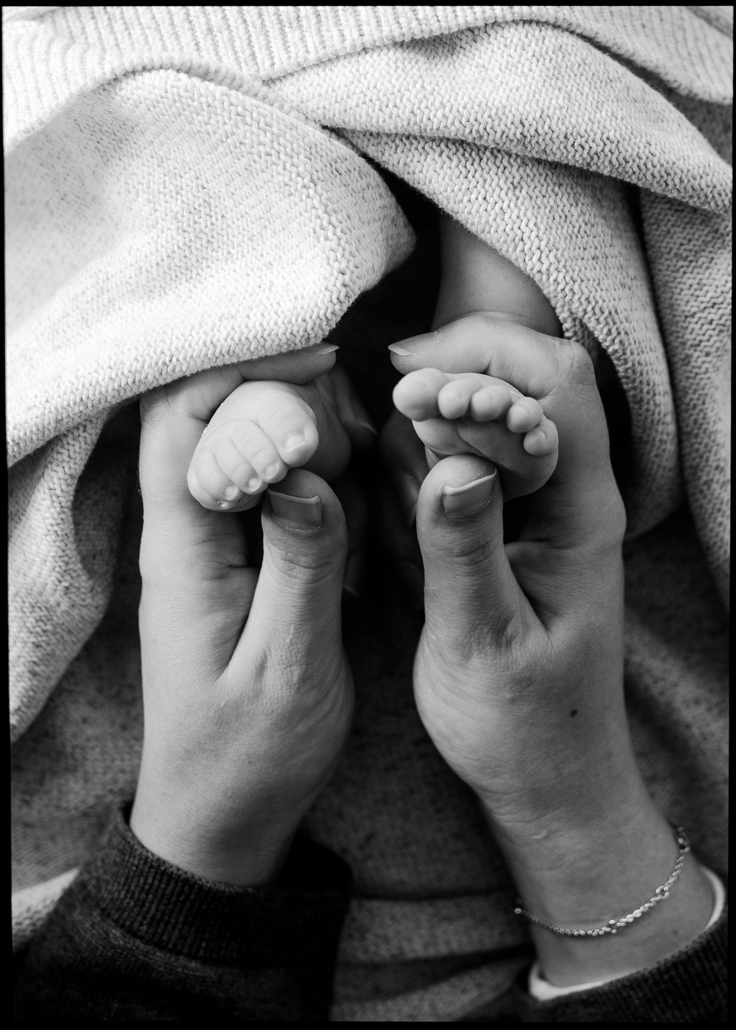 Black and white photo of a mother's hands gently holding a newborn baby's tiny feet, wrapped in a soft knit blanket.