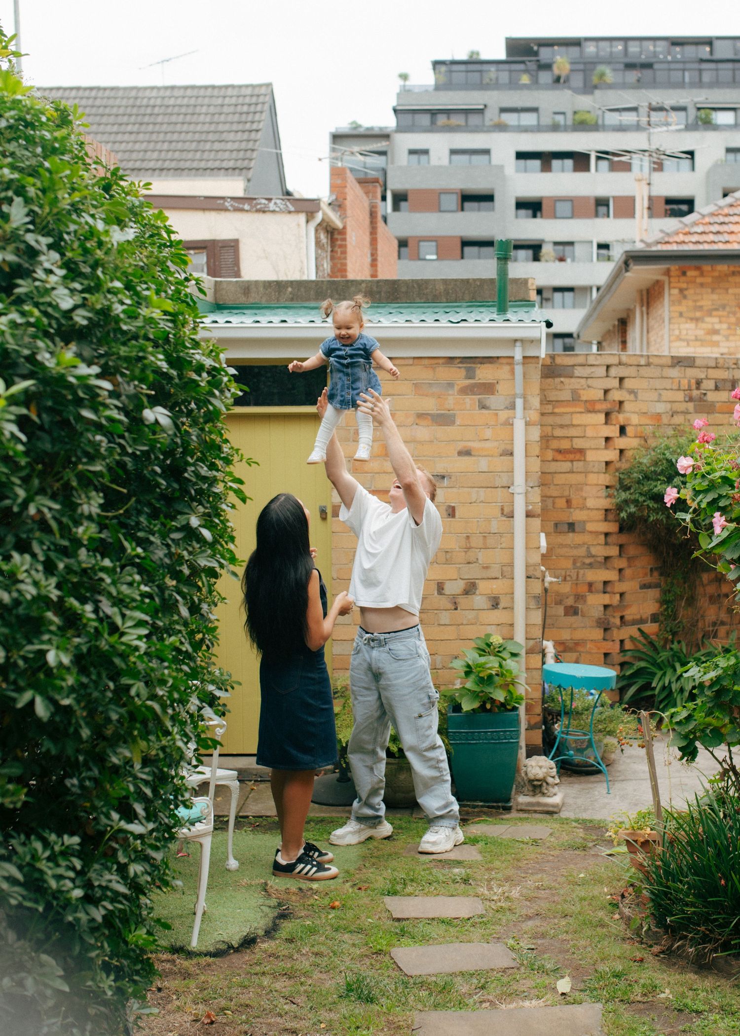 Father lifting smiling baby high in air in green urban backyard while mother watches.