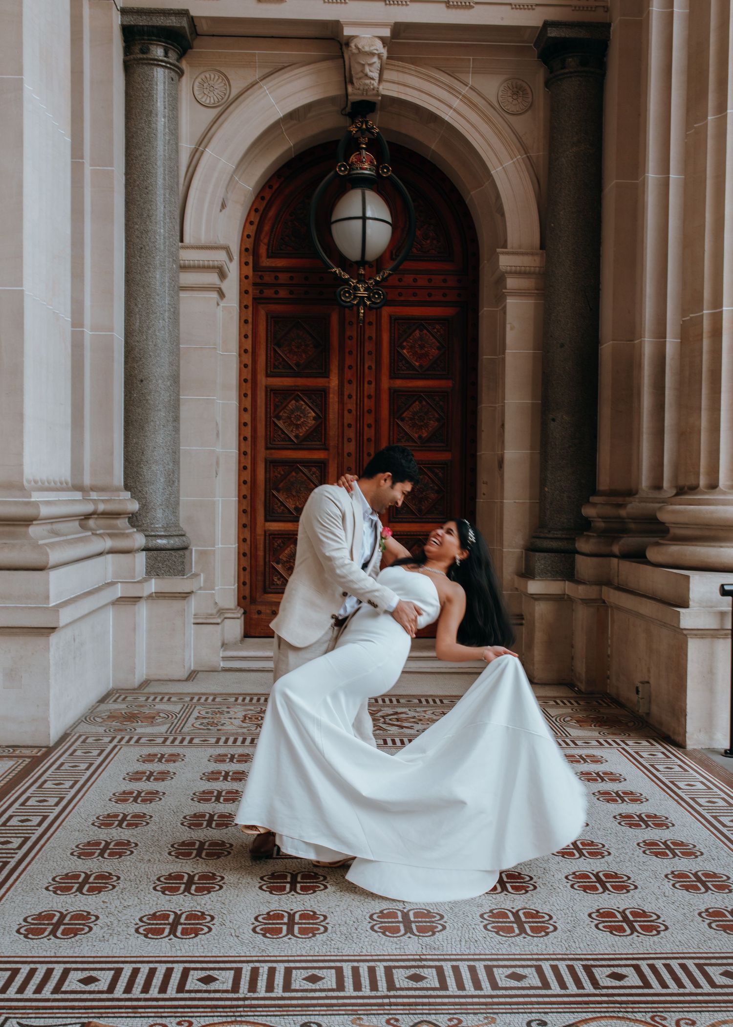 Groom dipping bride in elegant white gown before grand arched wooden door on mosaic floor.