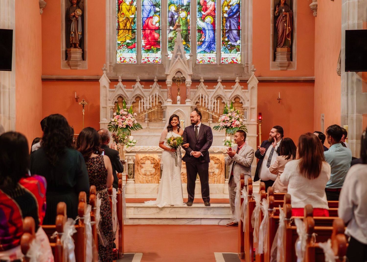Bride and groom stand at the altar of an elegant church adorned with floral arrangements and stained glass.
