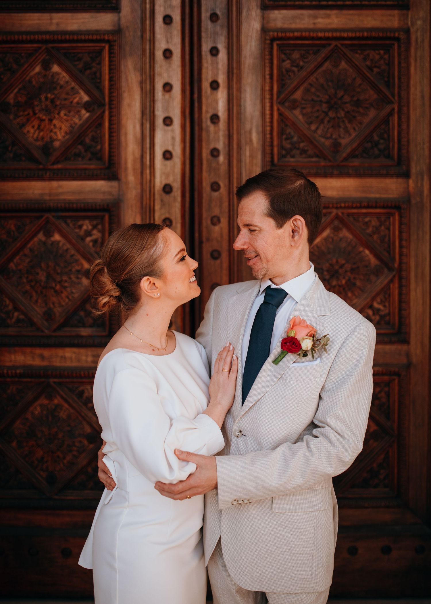 Elegant bride and groom gazing at each other before ornate carved wooden doors at wedding venue.
