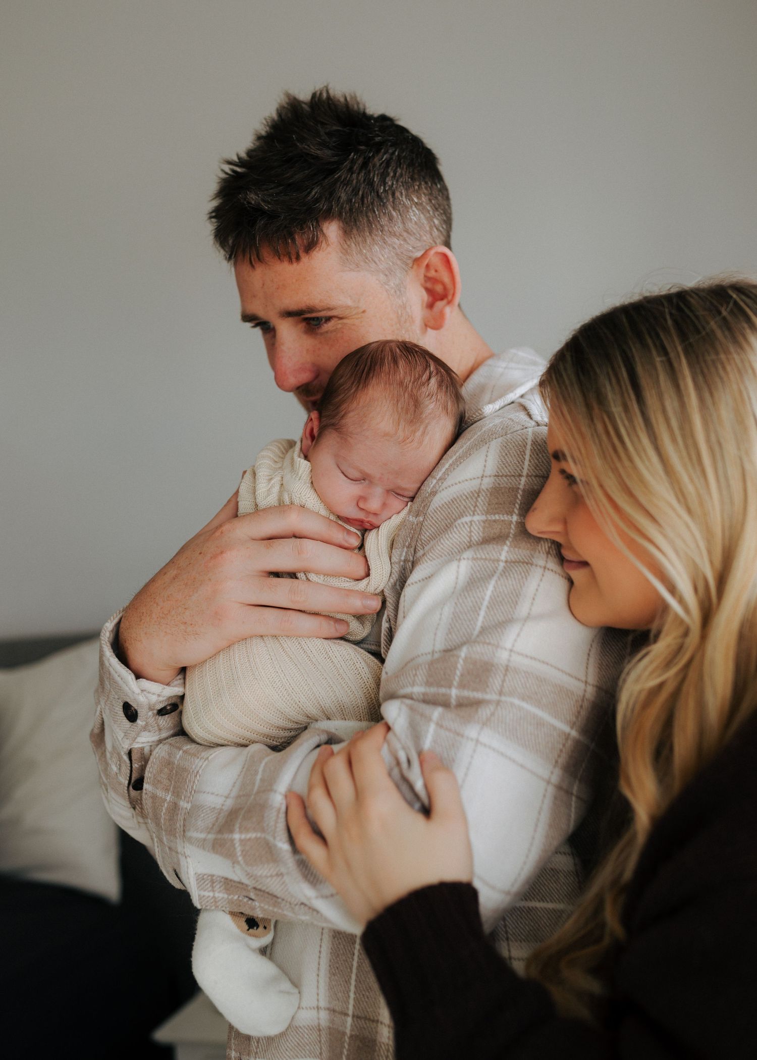 Father in plaid shirt holding sleeping newborn baby close to chest while mother gazes lovingly.