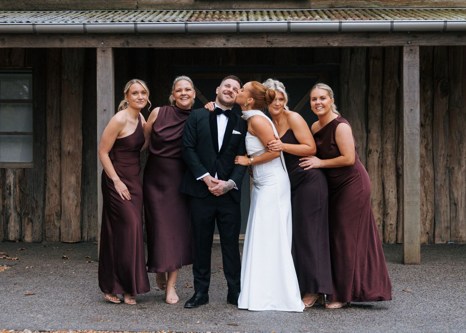 Bride and groom posing with four bridesmaids in burgundy gowns outside a rustic wooden building.
