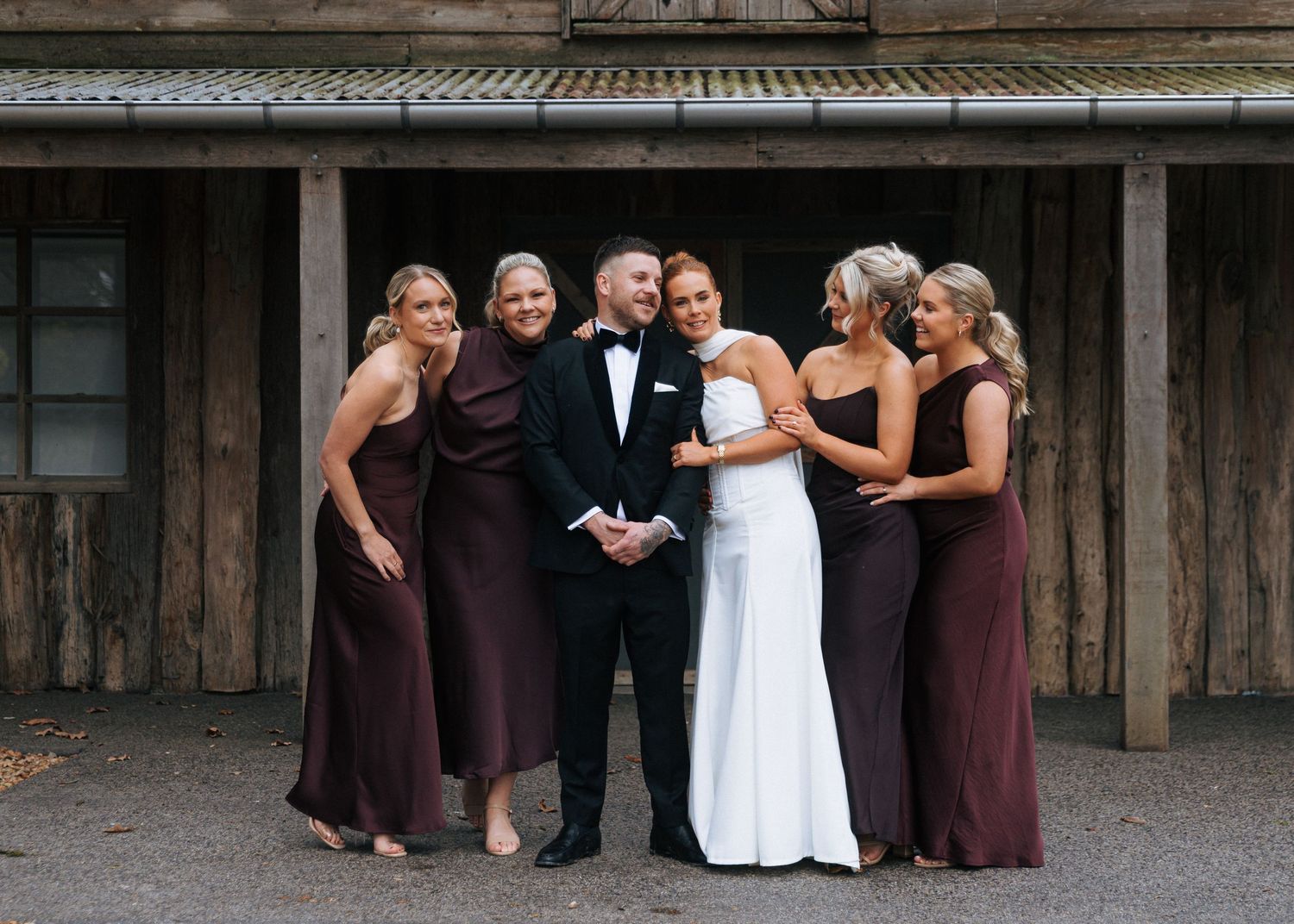 Groom in tuxedo poses with bride and four bridesmaids in burgundy gowns outside rustic wooden venue.
