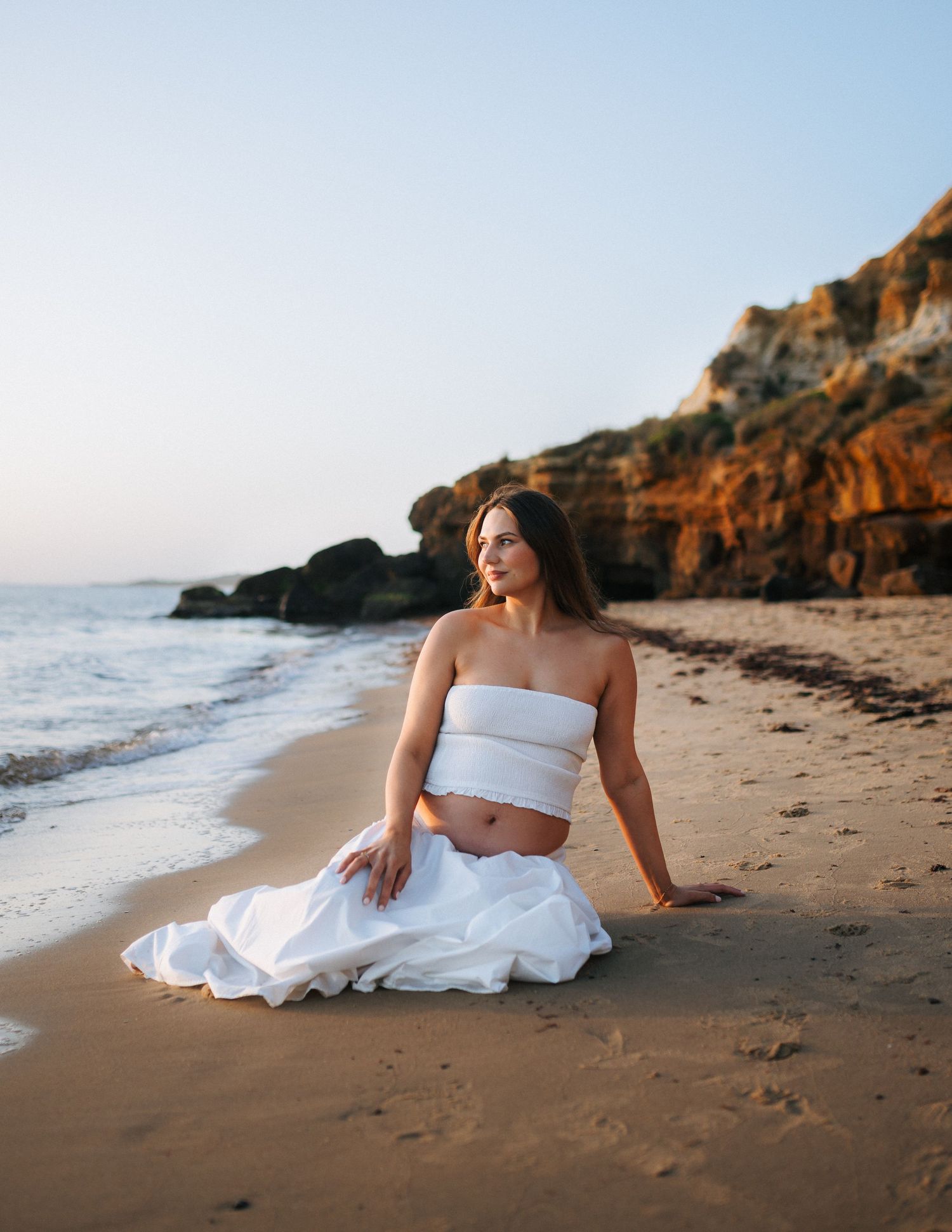 Pregnant woman in white two-piece outfit sits gracefully on sandy beach with rocky cliffs behind.
