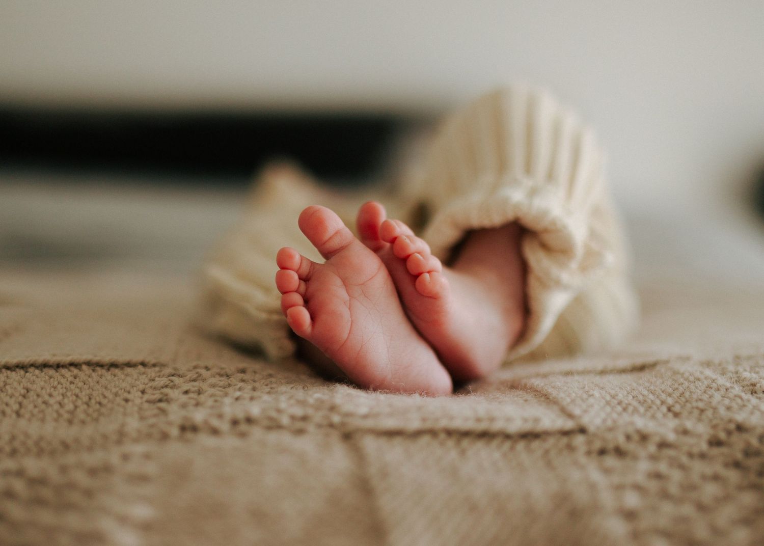 Close-up of tiny newborn baby feet peeking out from a cream knit sweater on a blanket.