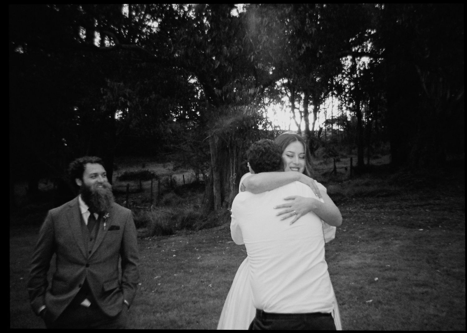 Black and white photo of bride hugging a guest warmly while bearded groom smiles nearby at outdoor wedding.