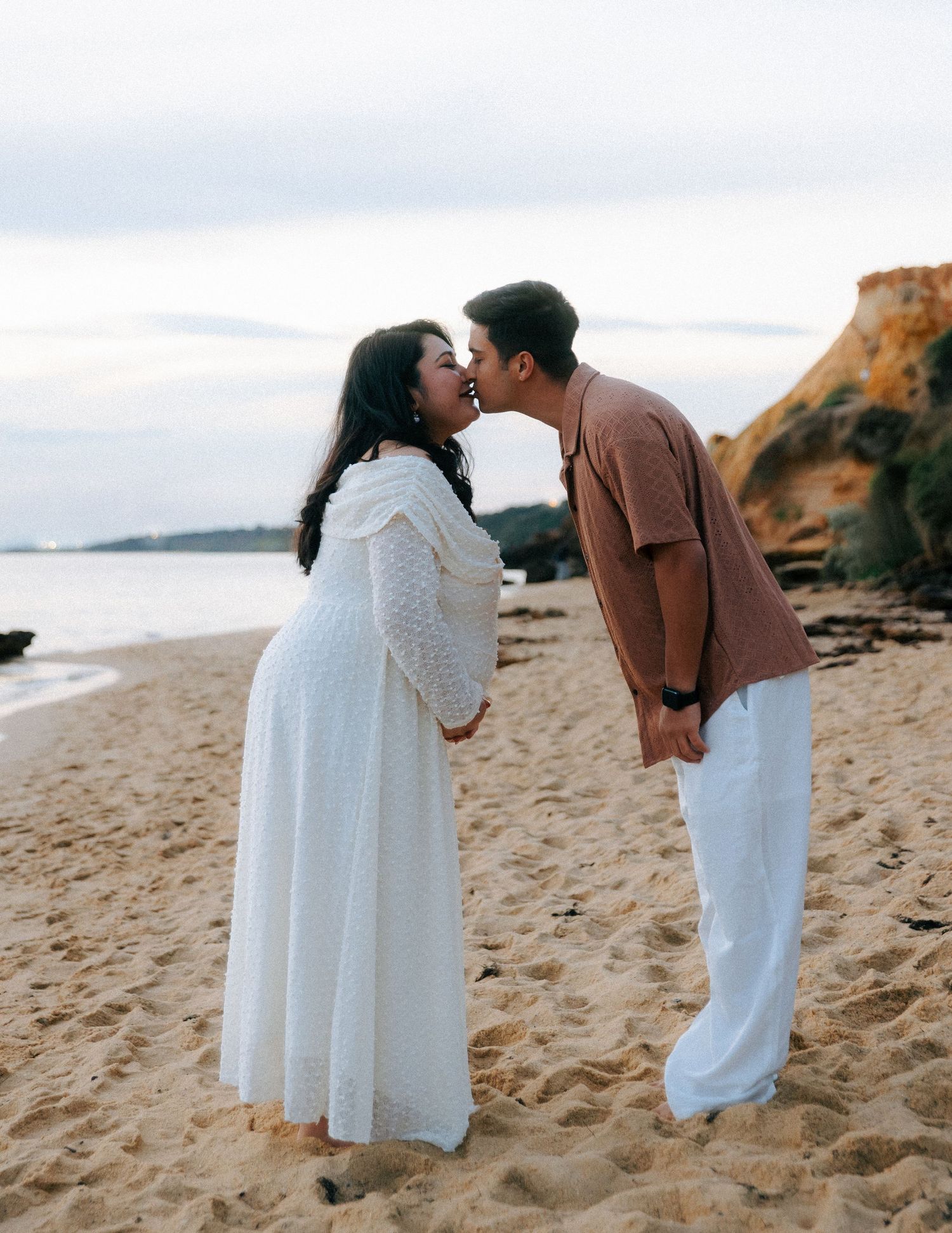 Pregnant couple sharing a kiss on a sandy beach at sunset.
