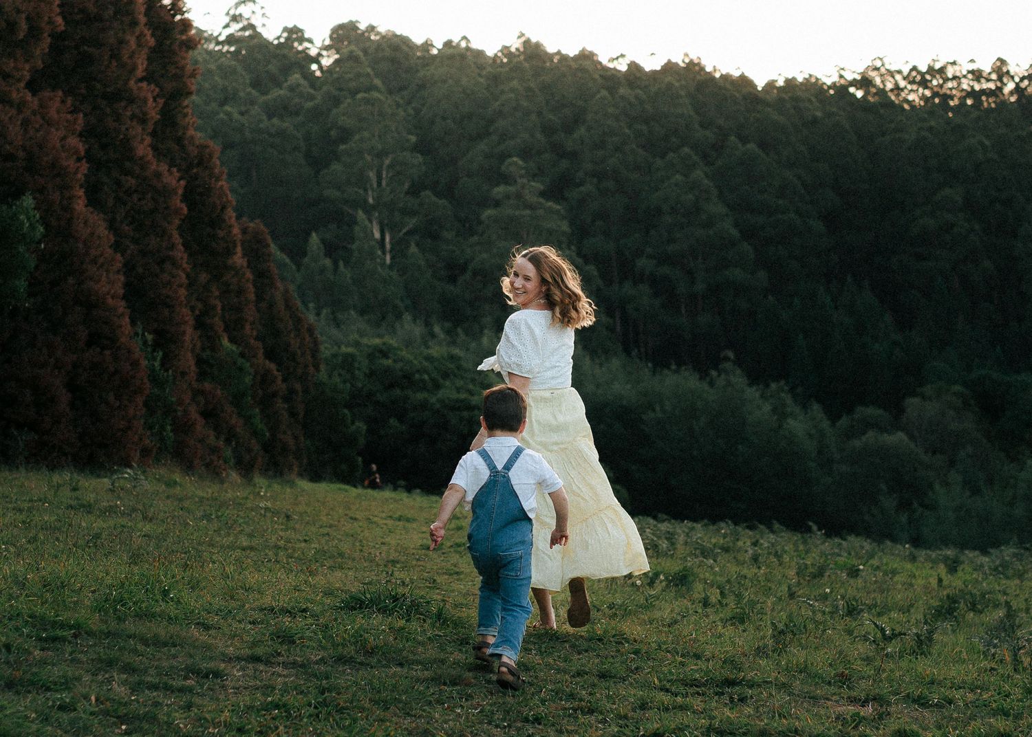 A mother in a white dress and her young son run together across a lush green hillside.