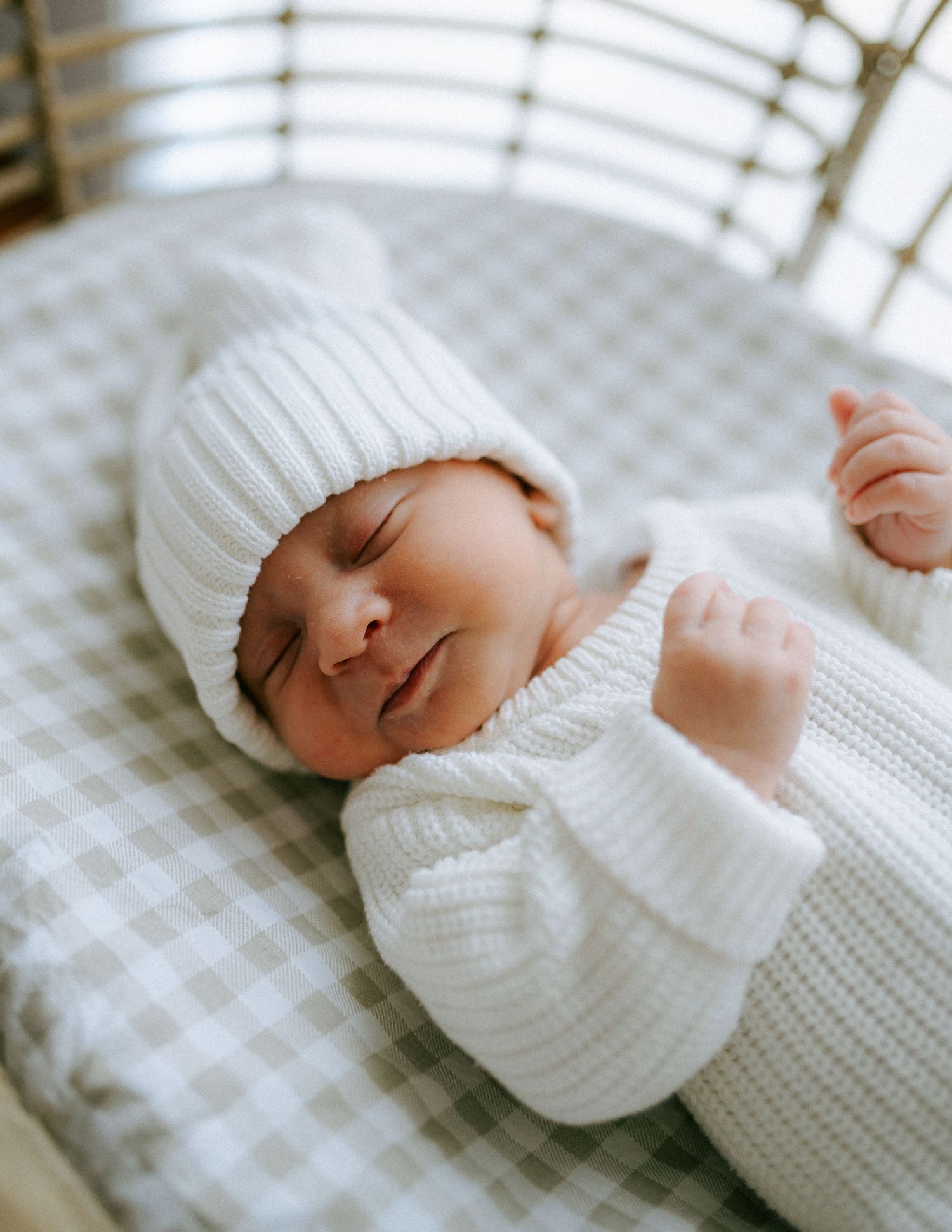 Close-up of peaceful sleeping newborn in white ribbed knit romper and beanie hat on gingham fabric.