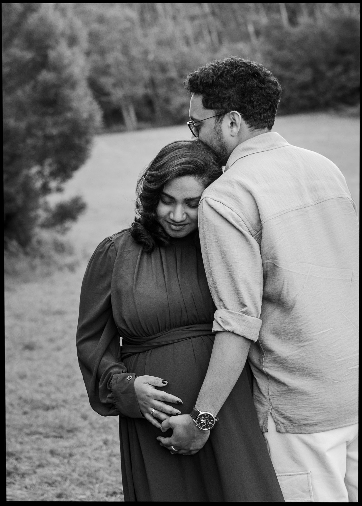 Black and white maternity photo of an expecting couple embracing outdoors as the man kisses her head and both hands rest on her baby bump.