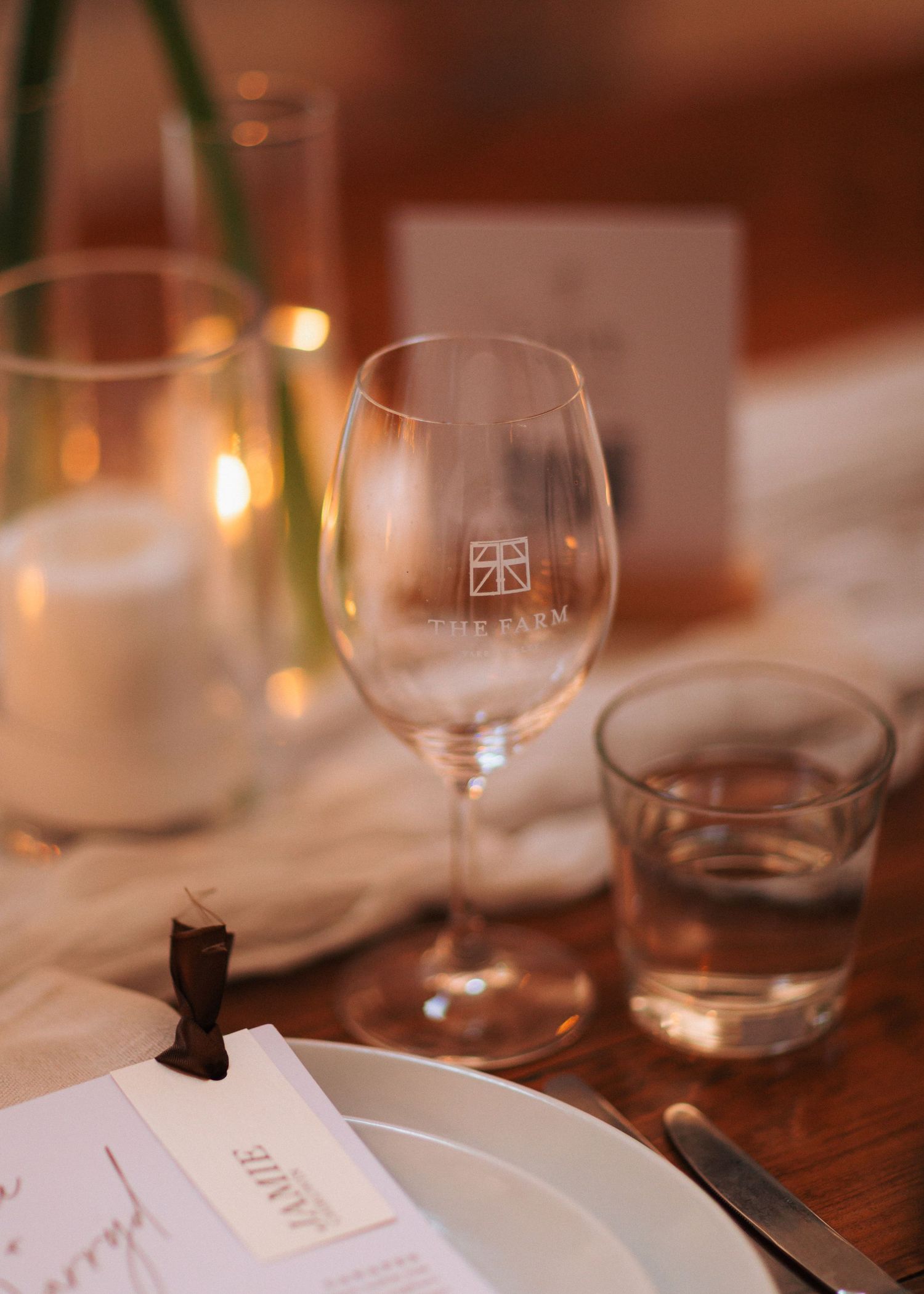 Elegant table setting with The Farm branded wine glass, candles, and place card on a wooden table.