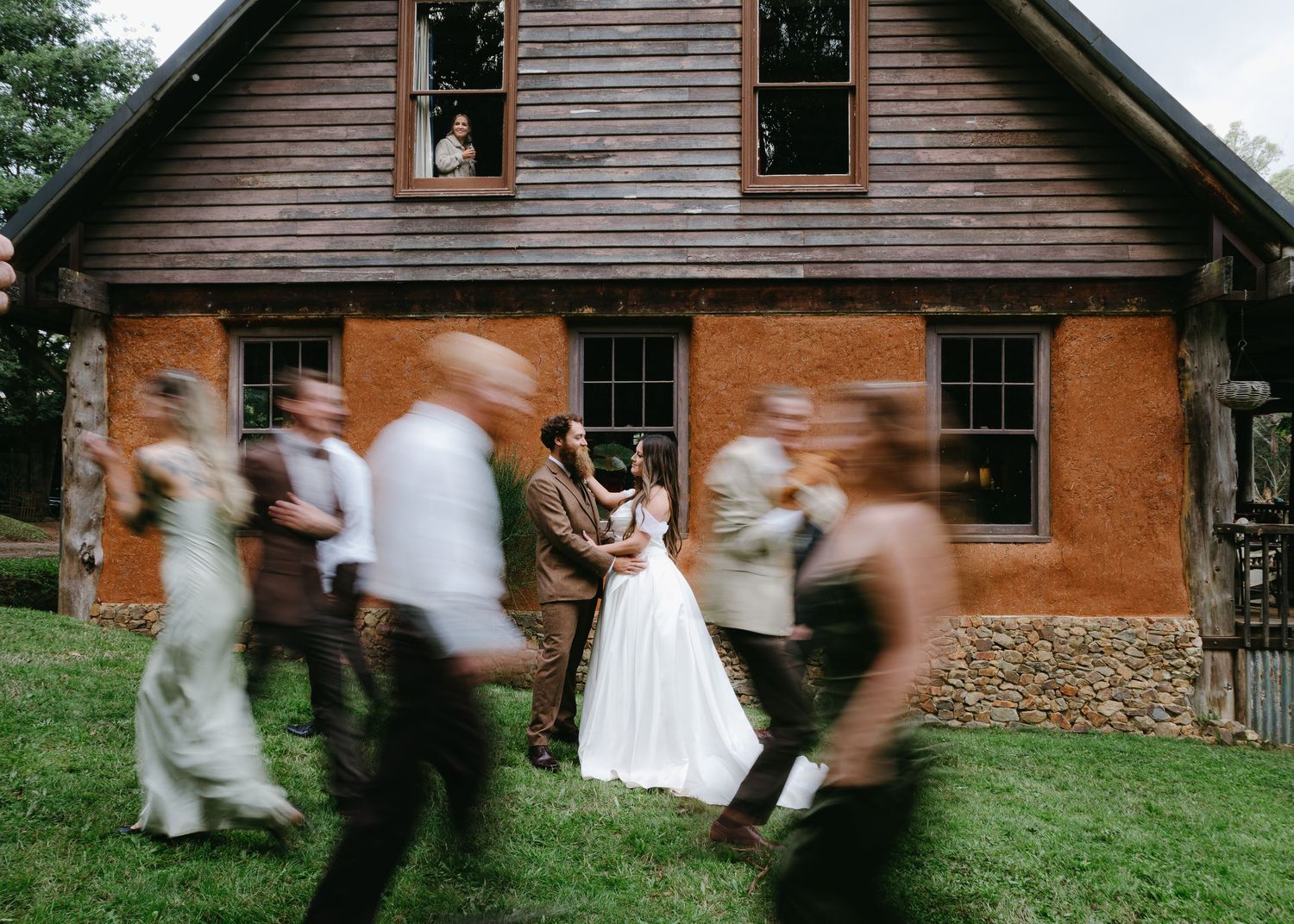 Bride and groom stand still as wedding party walks past rustic barn venue.