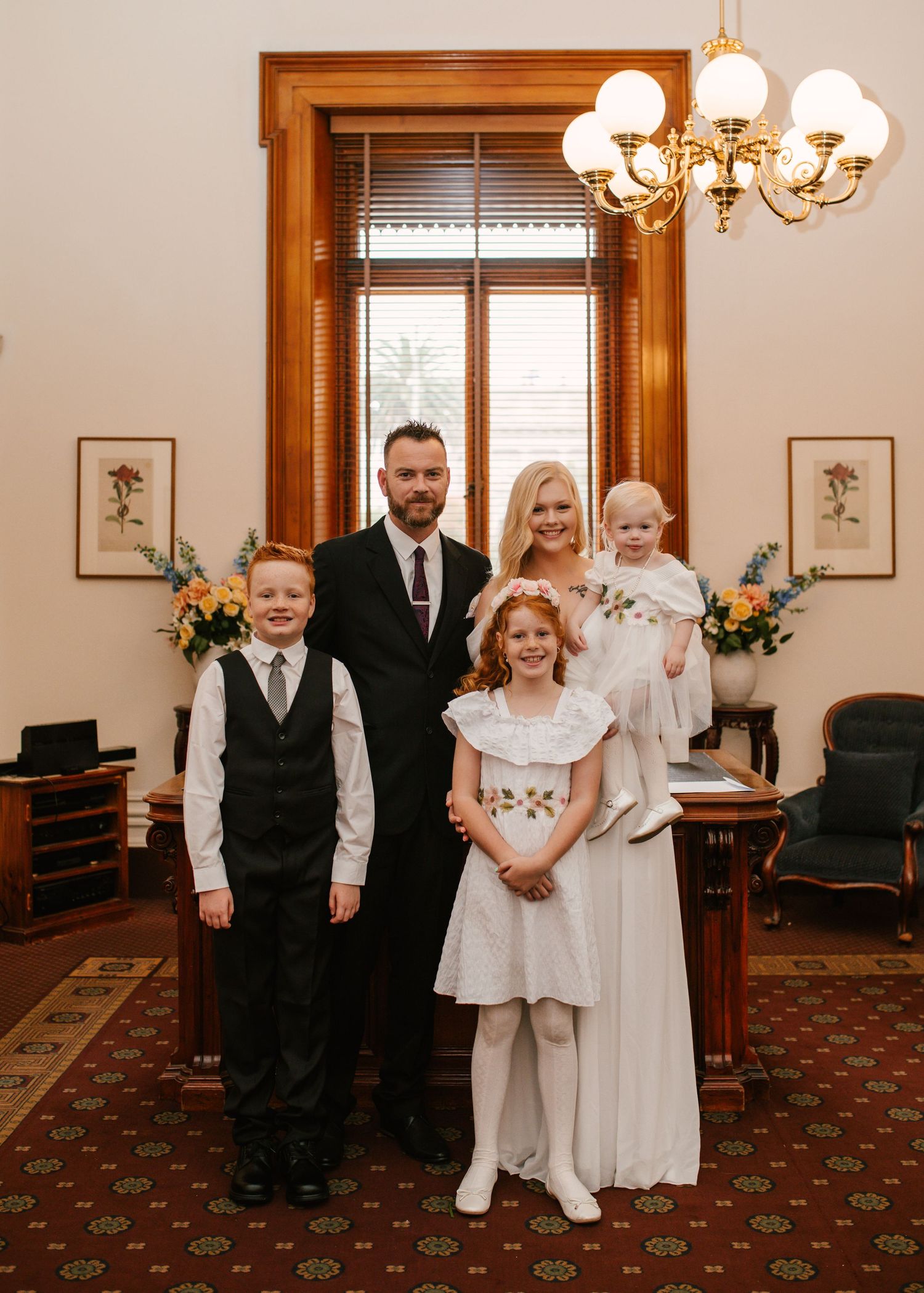 Family wedding portrait with bride, groom, and three children in formal attire inside registry office.