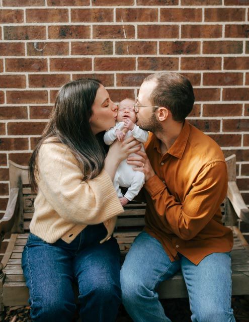 Parents kissing their newborn baby together while sitting on bench by brick wall.