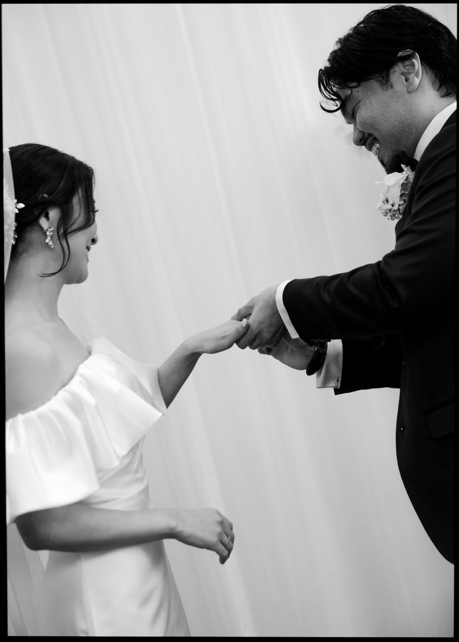 Black and white photo of smiling groom placing ring on bride's hand during wedding ceremony.