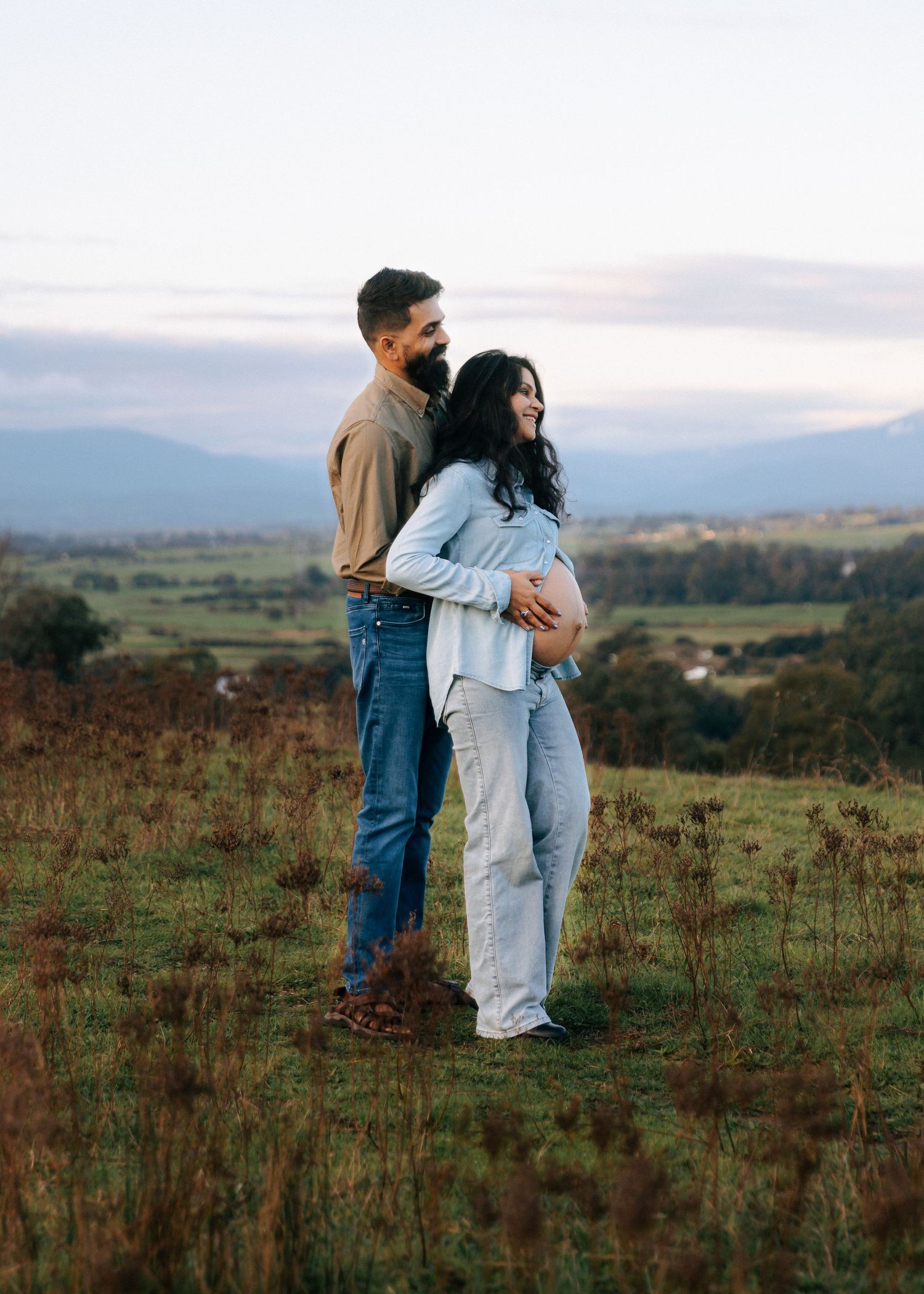 Couple embracing on a grassy hill at sunset, pregnant woman cradling her baby bump with scenic valley views.