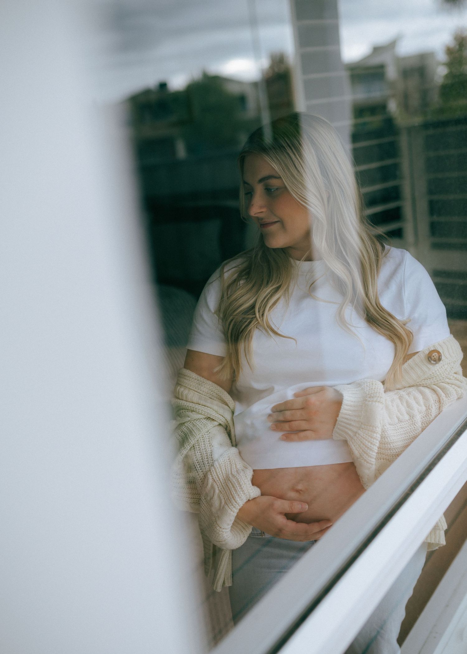 Pregnant woman in white crop top and beige cardigan holding bump, photographed through glass window.