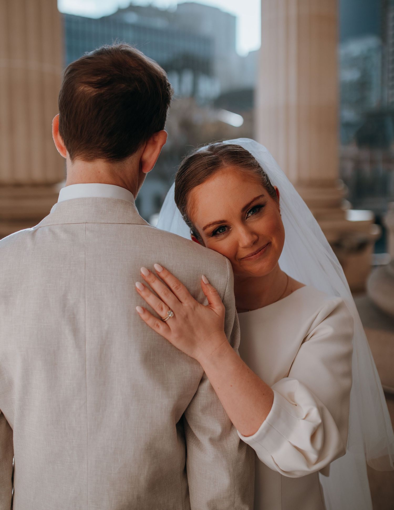 Smiling bride in white gown and veil resting head on groom's shoulder at outdoor wedding venue.