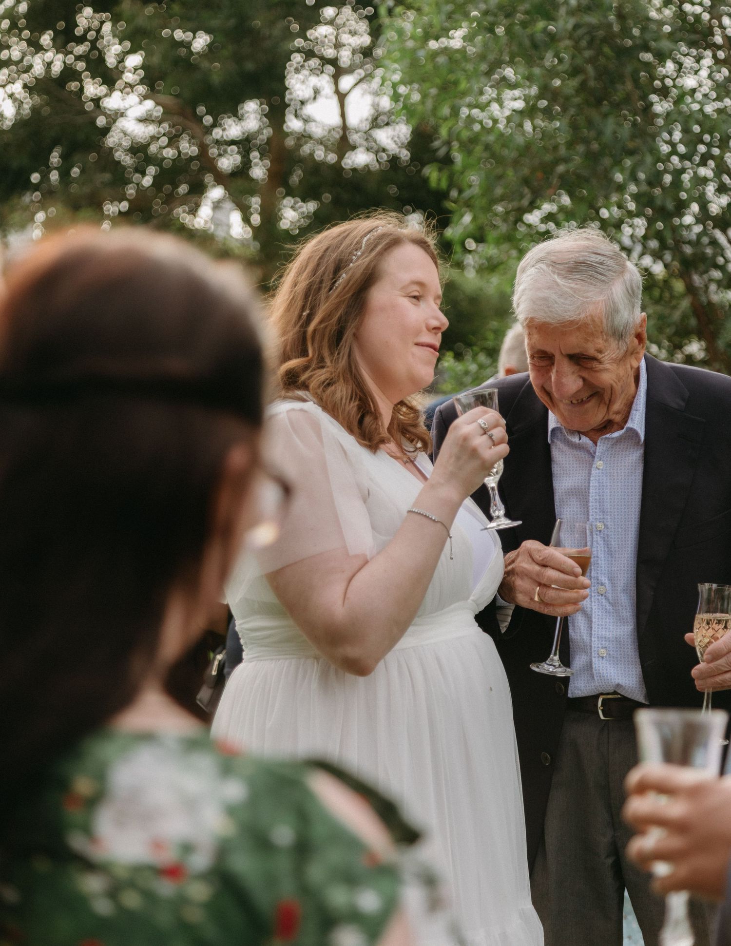 Bride in white dress laughing with elderly guest while holding champagne at outdoor wedding reception.