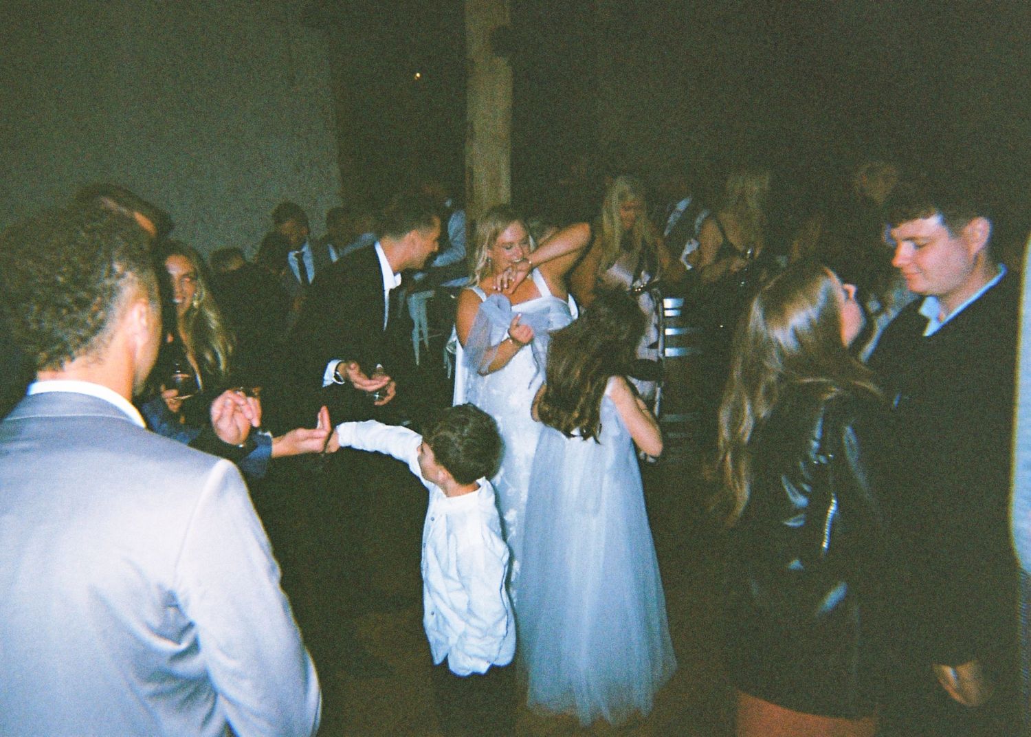 Wedding guests including a bride in a blue gown dance together on a dimly lit dance floor at a reception.