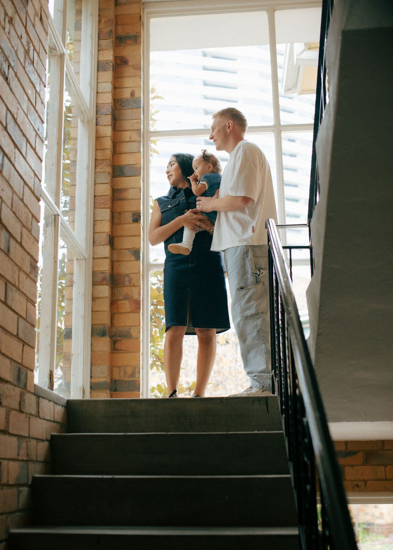 Family with baby standing at top of stairs by large window in brick home interior.