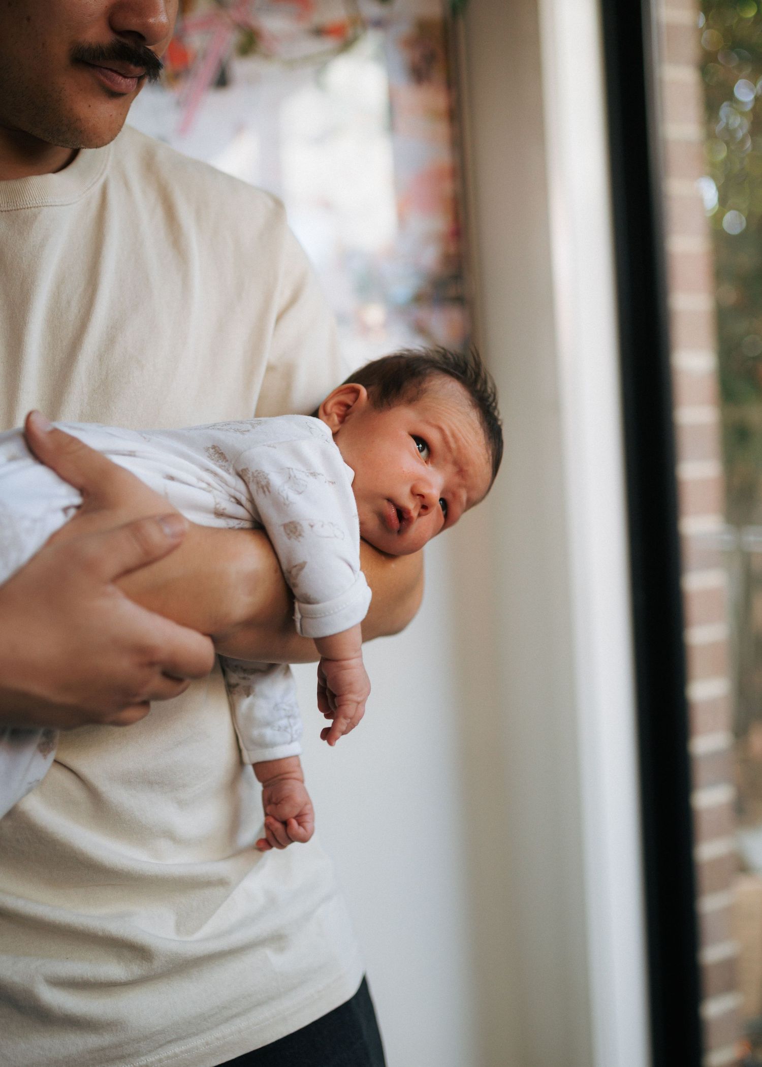 A father in a cream t-shirt gently cradles a wide-eyed newborn baby in his arms beside a bright window.