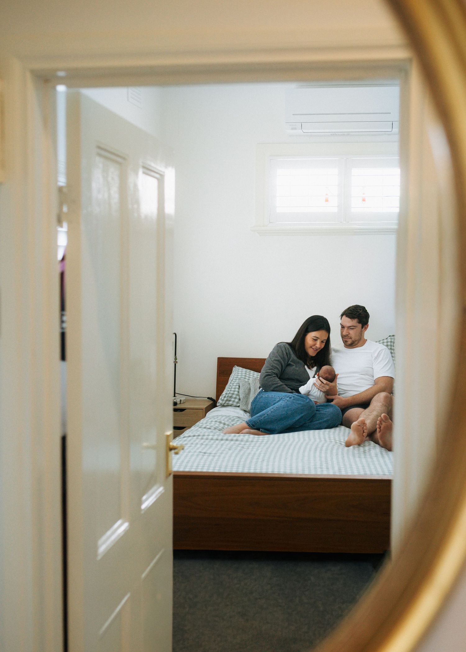 Mirror reflection of a smiling couple sitting on a bed, cradling their newborn baby in a bright, cozy bedroom.