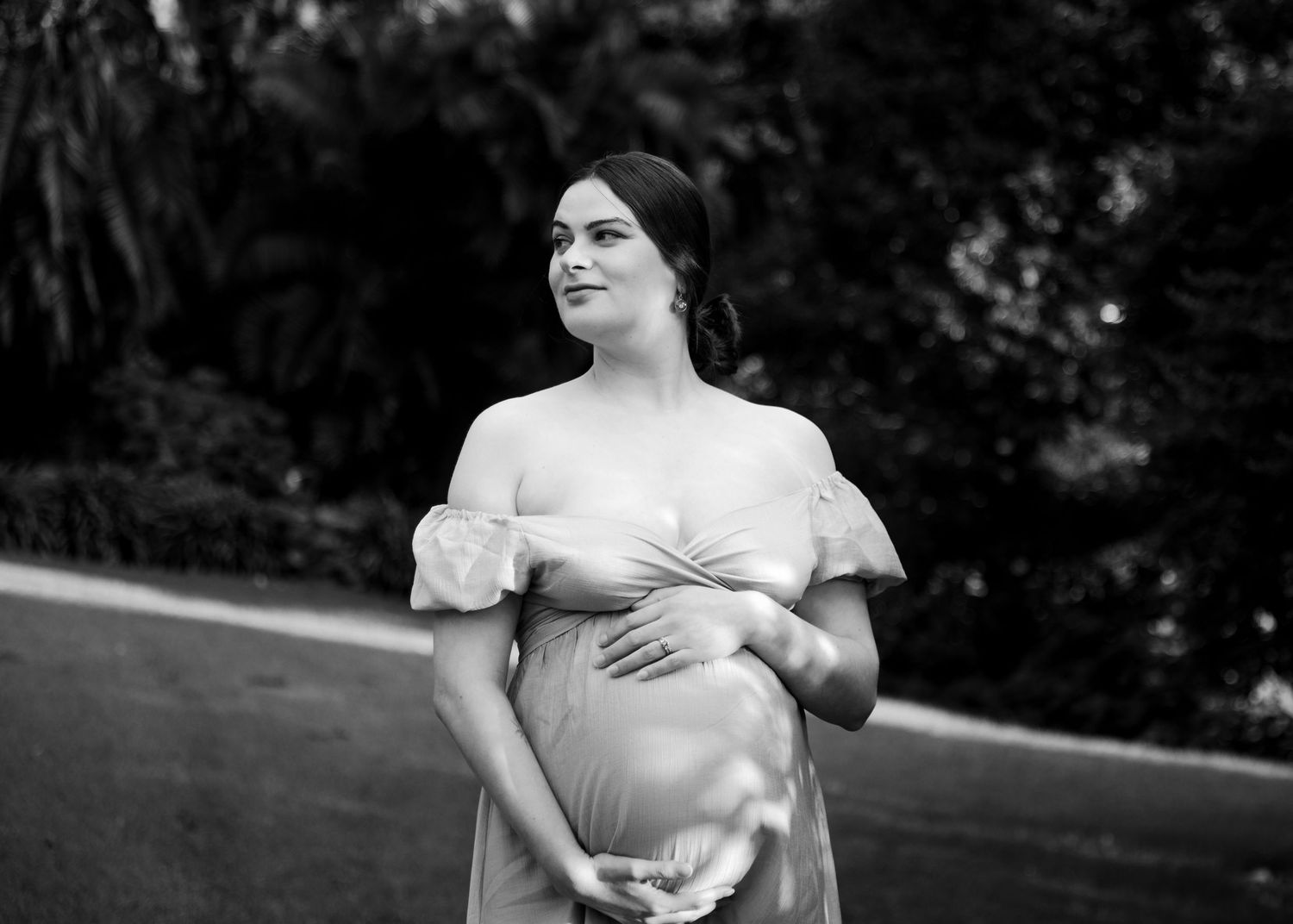 Black and white maternity portrait of pregnant woman in off-shoulder gown cradling belly in lush garden.
