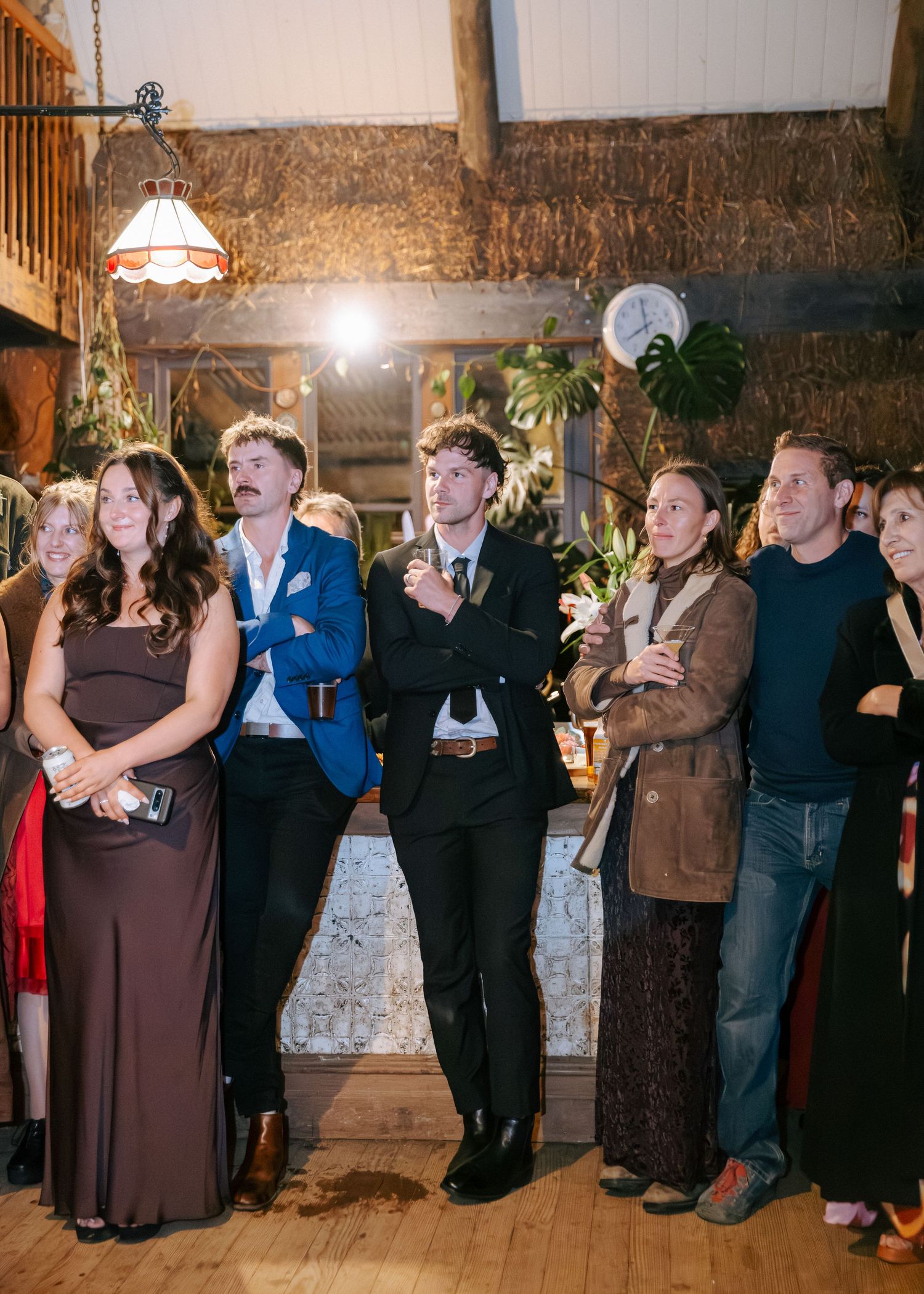Wedding guests gather indoors in a rustic barn venue, dressed in semi-formal attire, watching a ceremony.