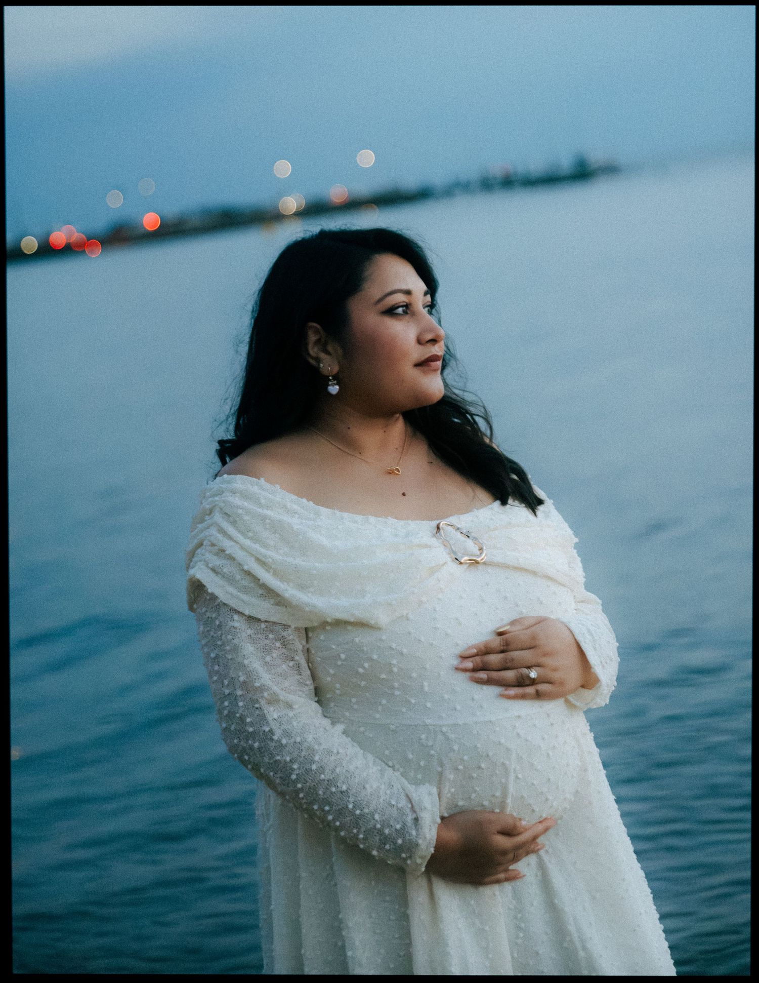 Pregnant woman in white beaded off-shoulder dress holding belly near ocean at twilight.