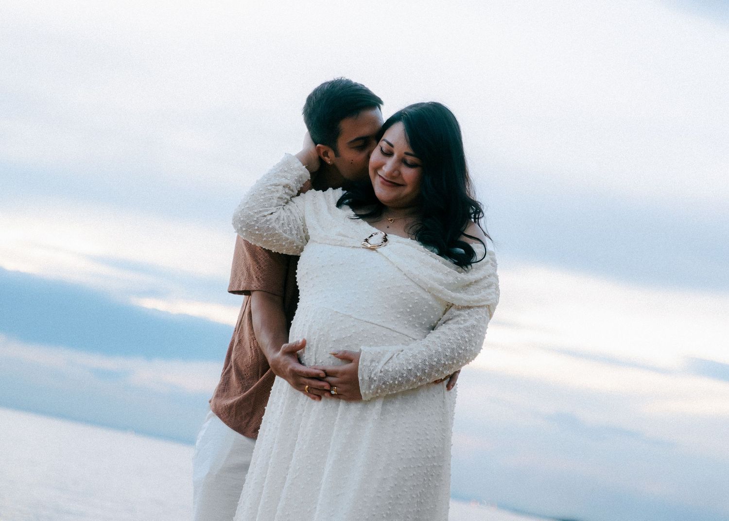 Romantic maternity photo of couple embracing by the ocean, woman in elegant white lace dress.
