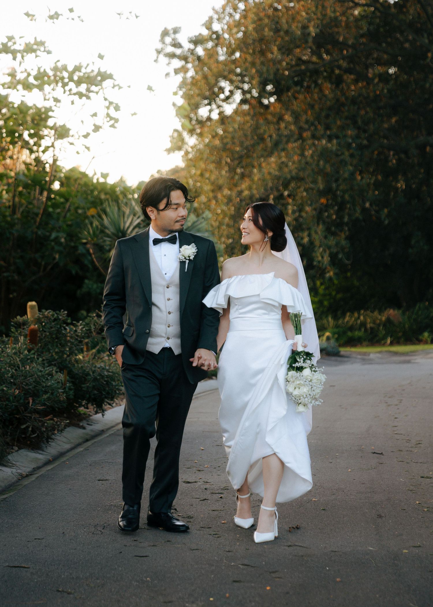 Smiling bride and groom in wedding attire walking hand in hand along a sunlit garden path at golden hour.